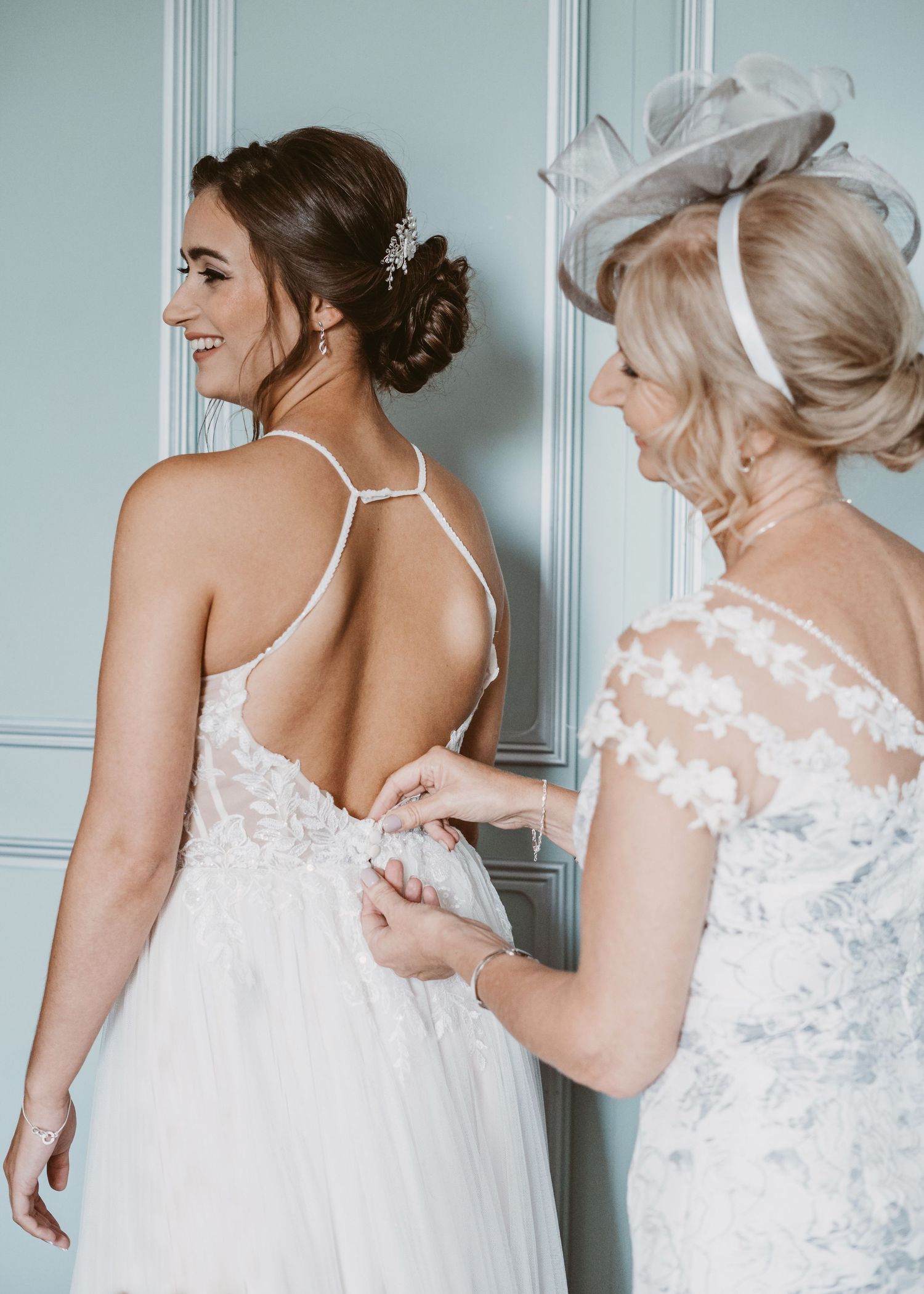 Two women in white dresses prepare for a wedding, adjusting the back of an elegant gown.