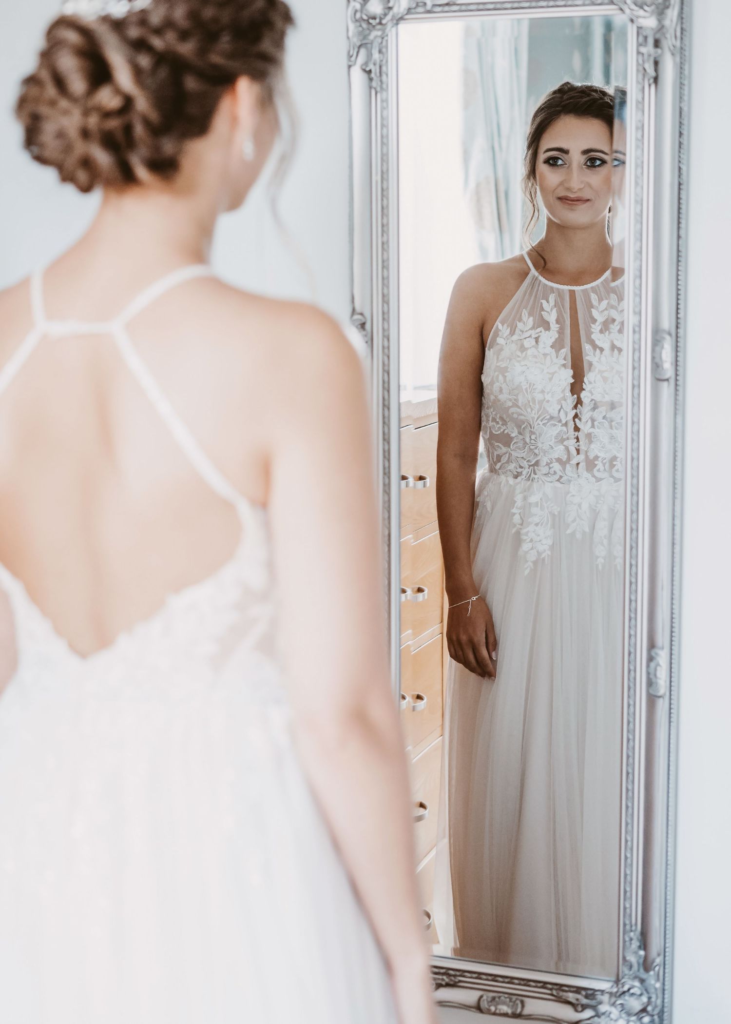 A bride admires her reflection in an ornate vintage mirror while wearing a delicate lace wedding gown.