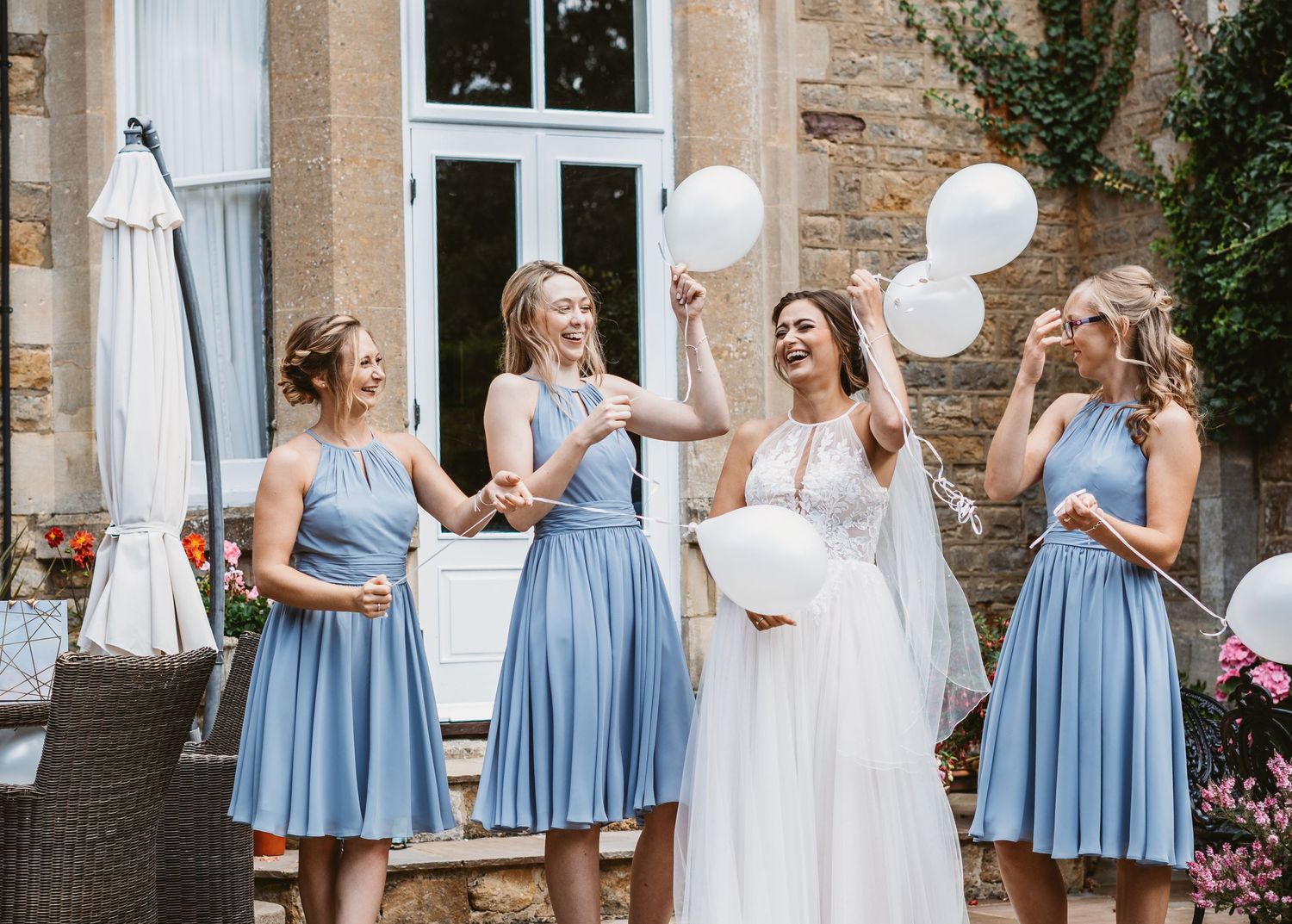 Bridesmaids in light blue dresses celebrate with white balloons outdoors.