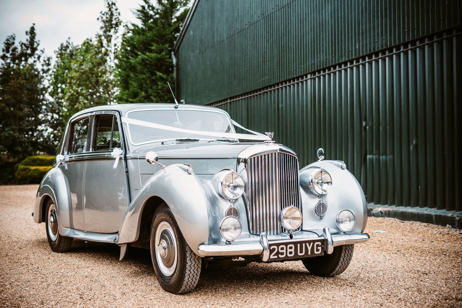 A classic silver Bentley vintage car parked outside a green industrial building.