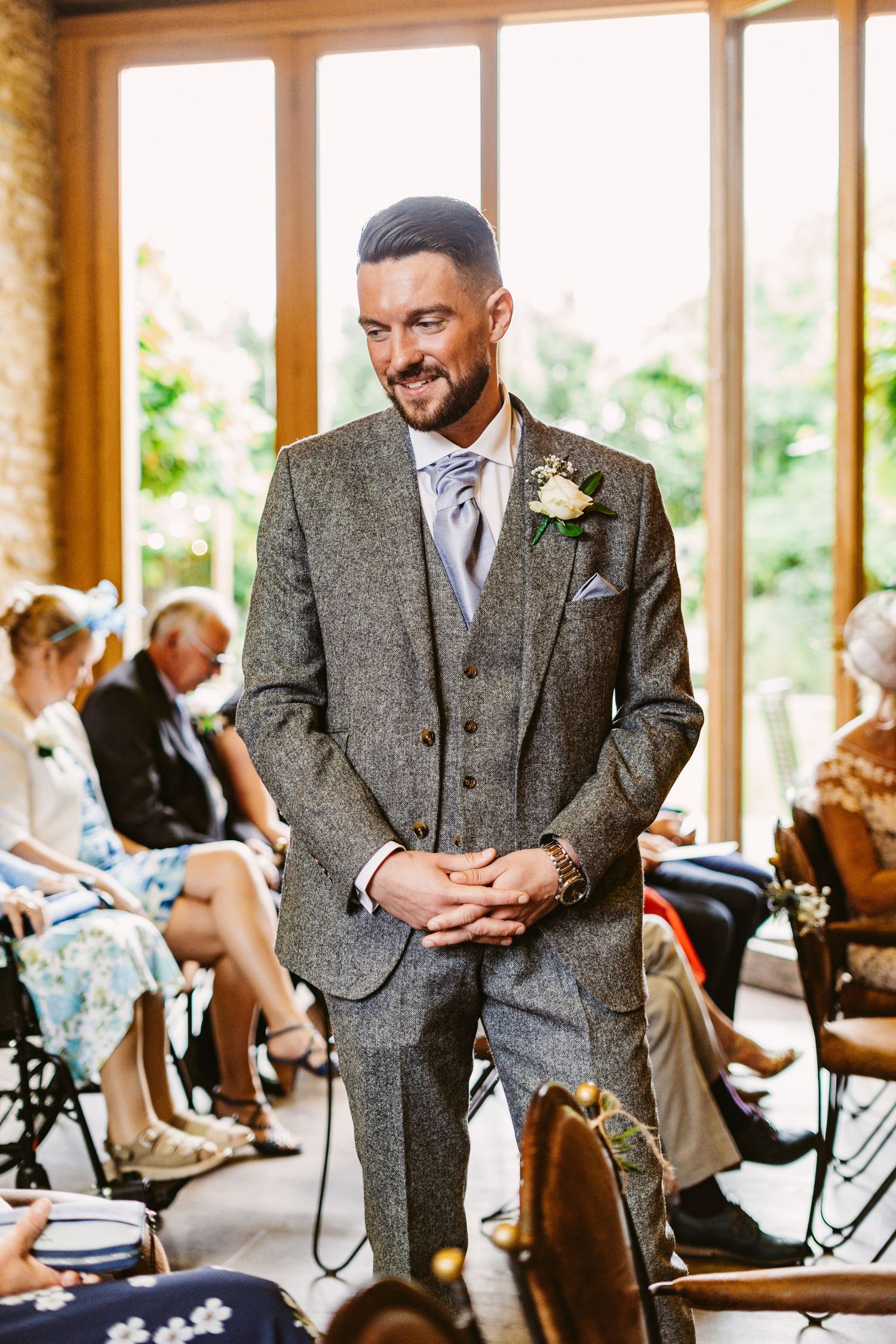 A groom in a grey tweed suit smiles while standing during a wedding ceremony in a rustic venue.