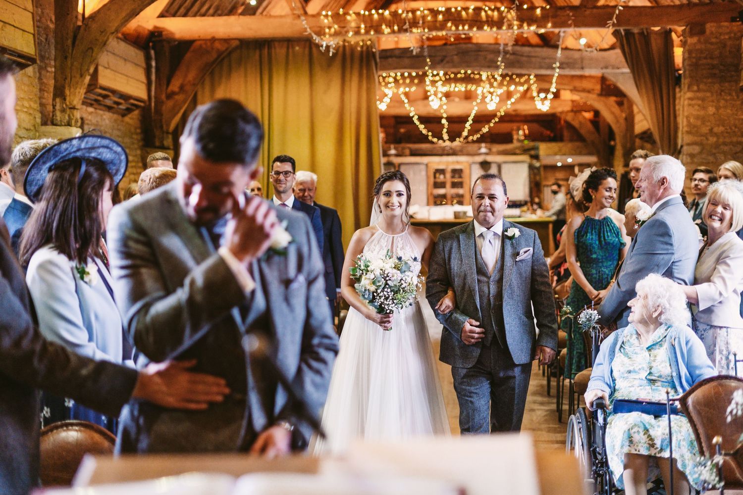Guests watch a wedding ceremony in a rustic barn venue with wooden beams and chandeliers.