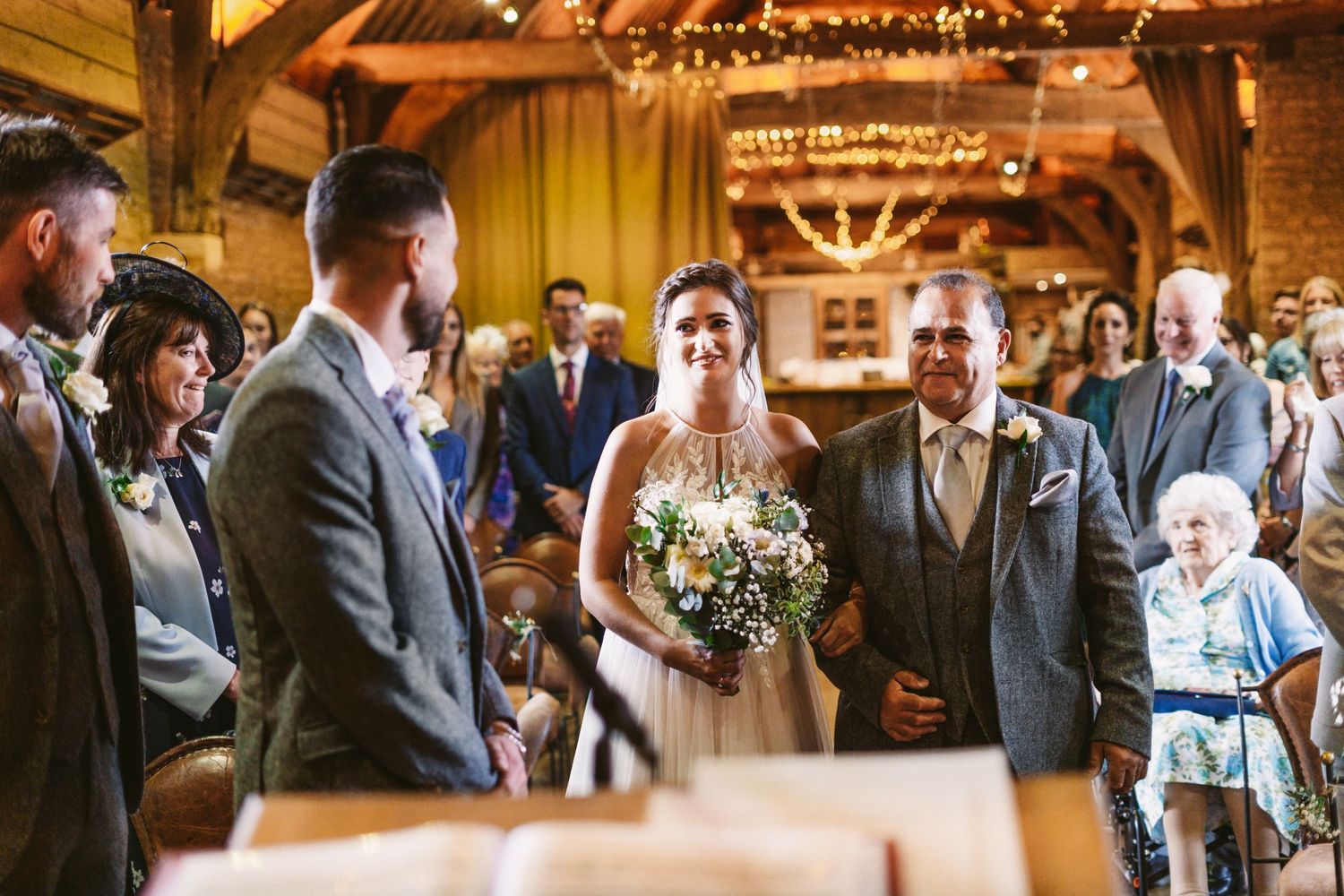 A wedding ceremony takes place in a beautiful rustic barn with exposed beams and twinkling lights.