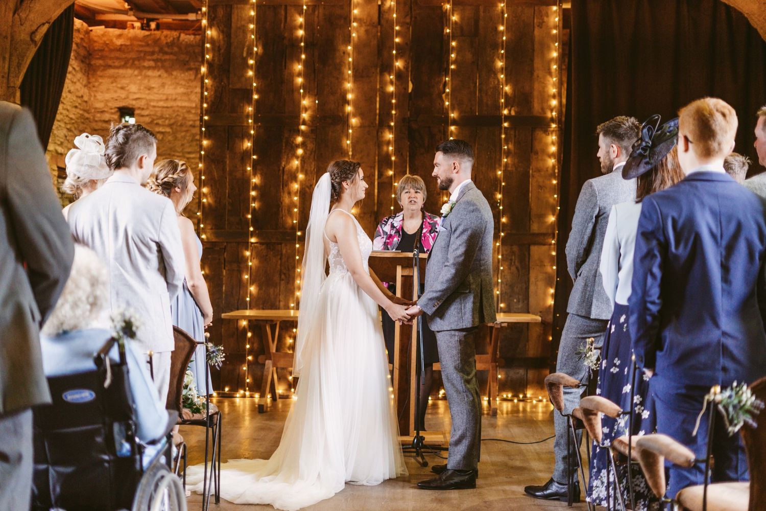 Wedding ceremony under string lights in rustic brick venue with guests seated.