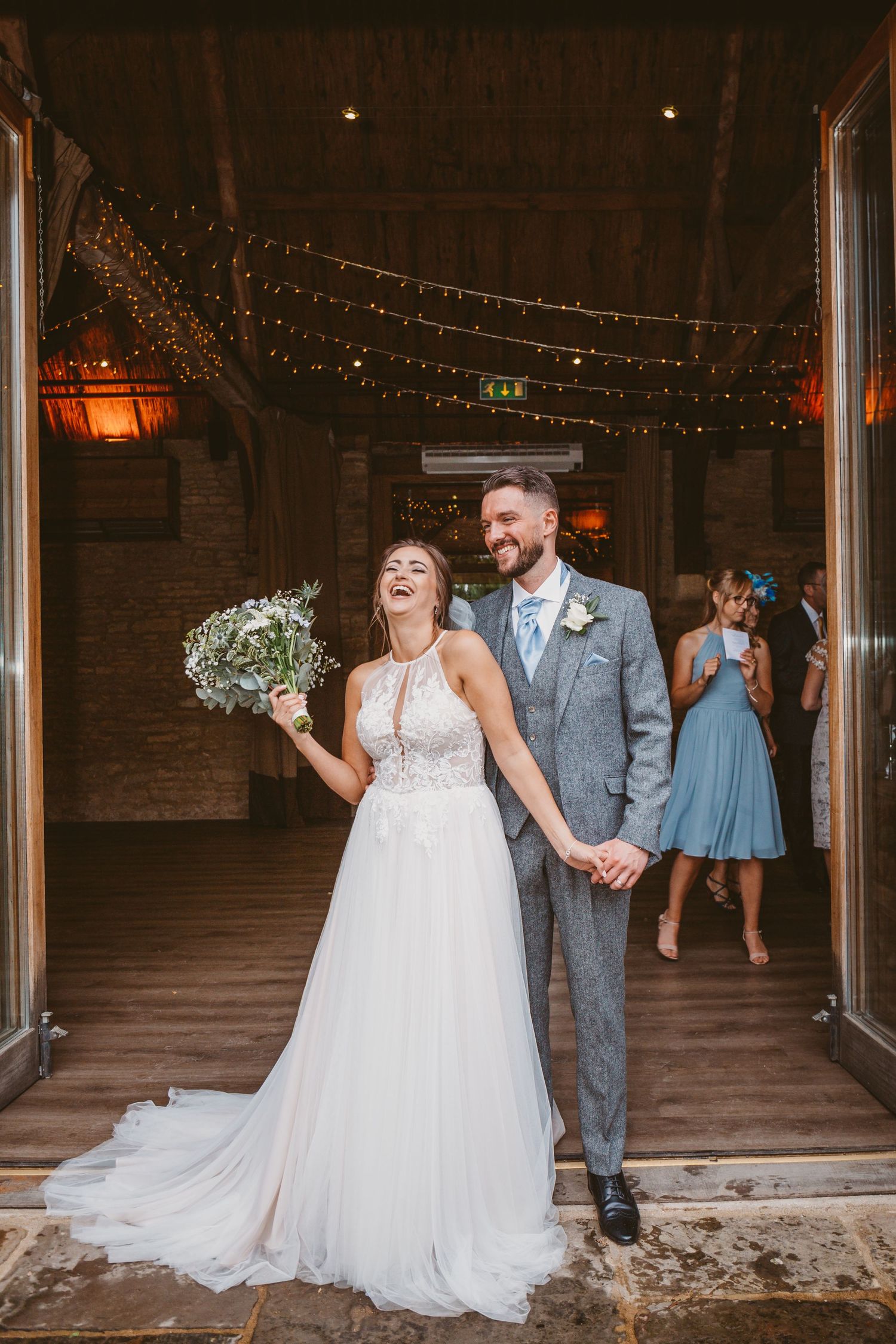 A joyous wedding couple laughs together under twinkling lights at a rustic venue entrance.