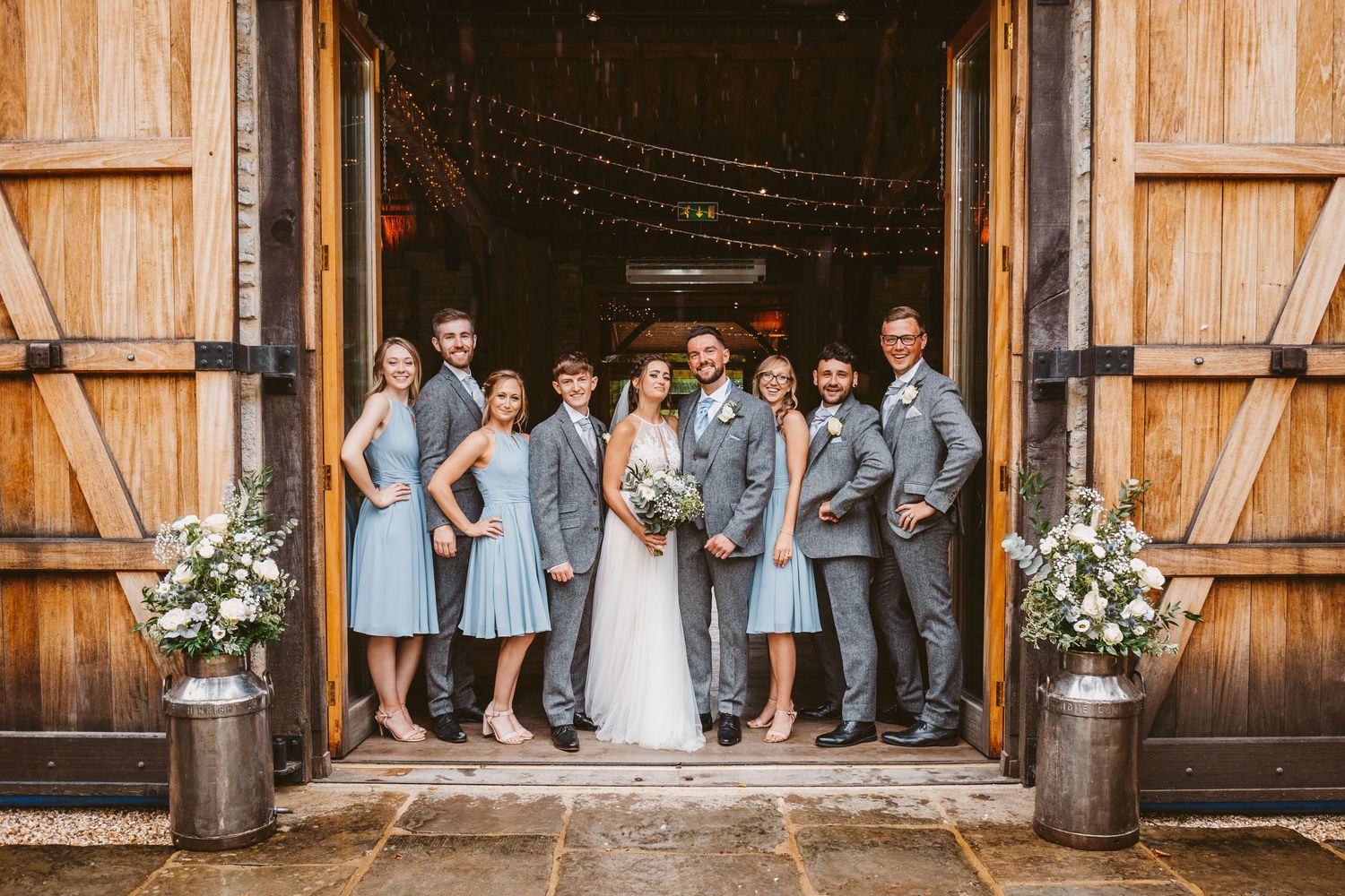 Wedding party poses in rustic barn doorway with bridesmaids in blue dresses and groomsmen in gray suits.