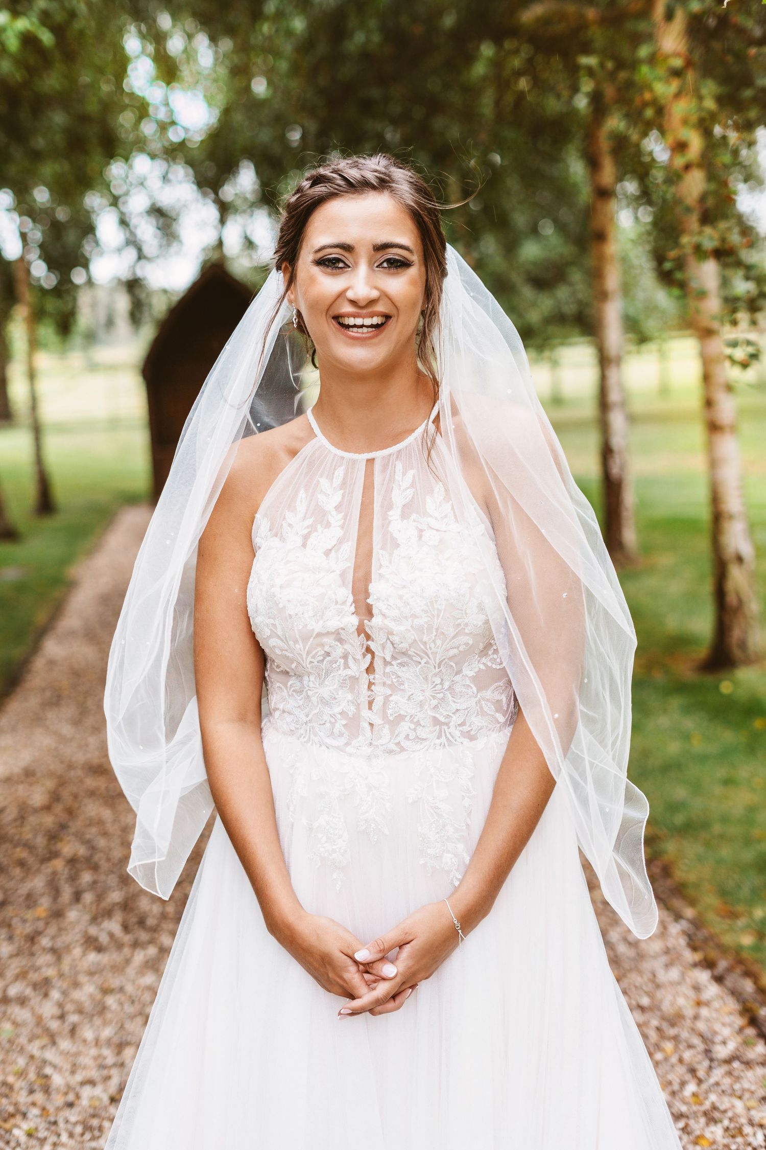 Bride in halter-style lace wedding gown with flowing veil stands on garden path.