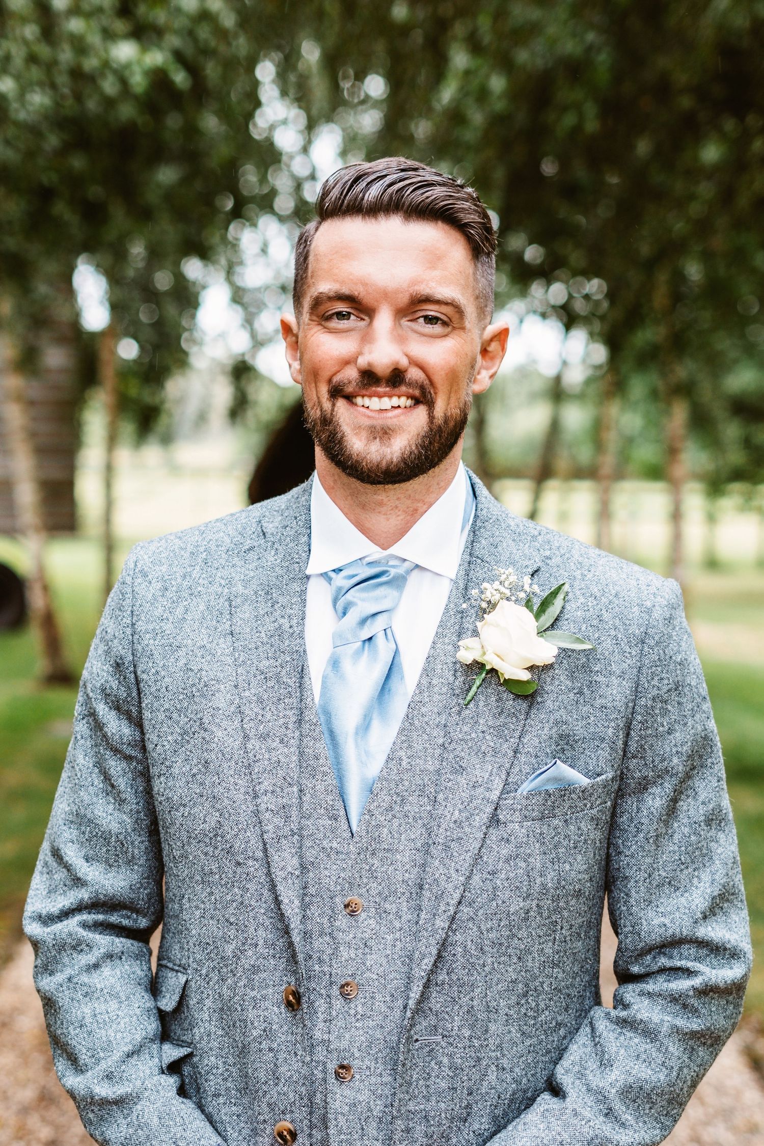 Groom in gray suit with light blue tie and white boutonniere smiles outdoors.