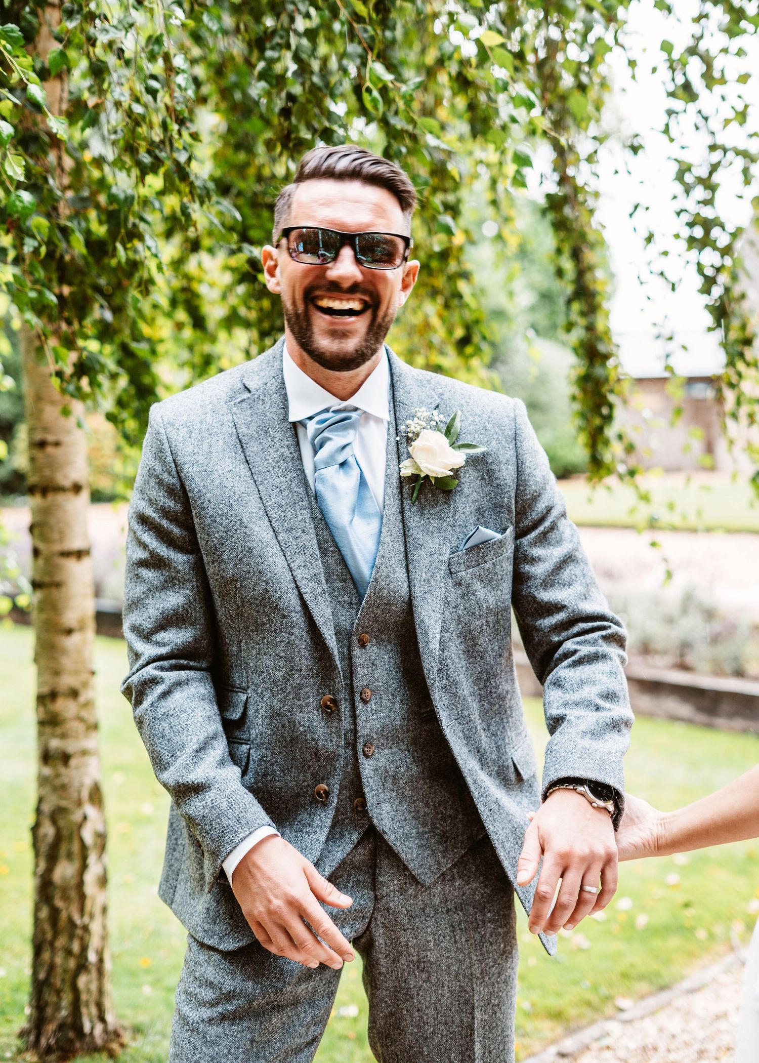 Groom in sunglasses and gray suit laughs joyfully in garden setting.