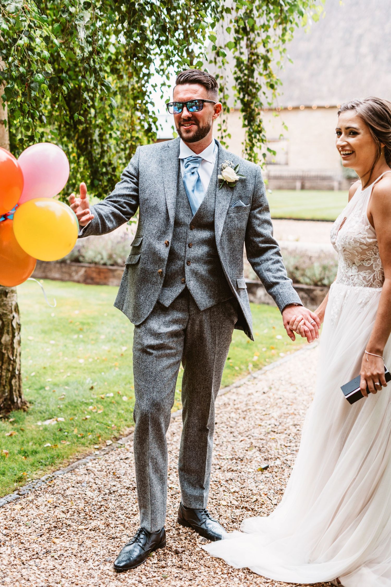 Couple holds colorful balloons while walking down garden path at wedding.