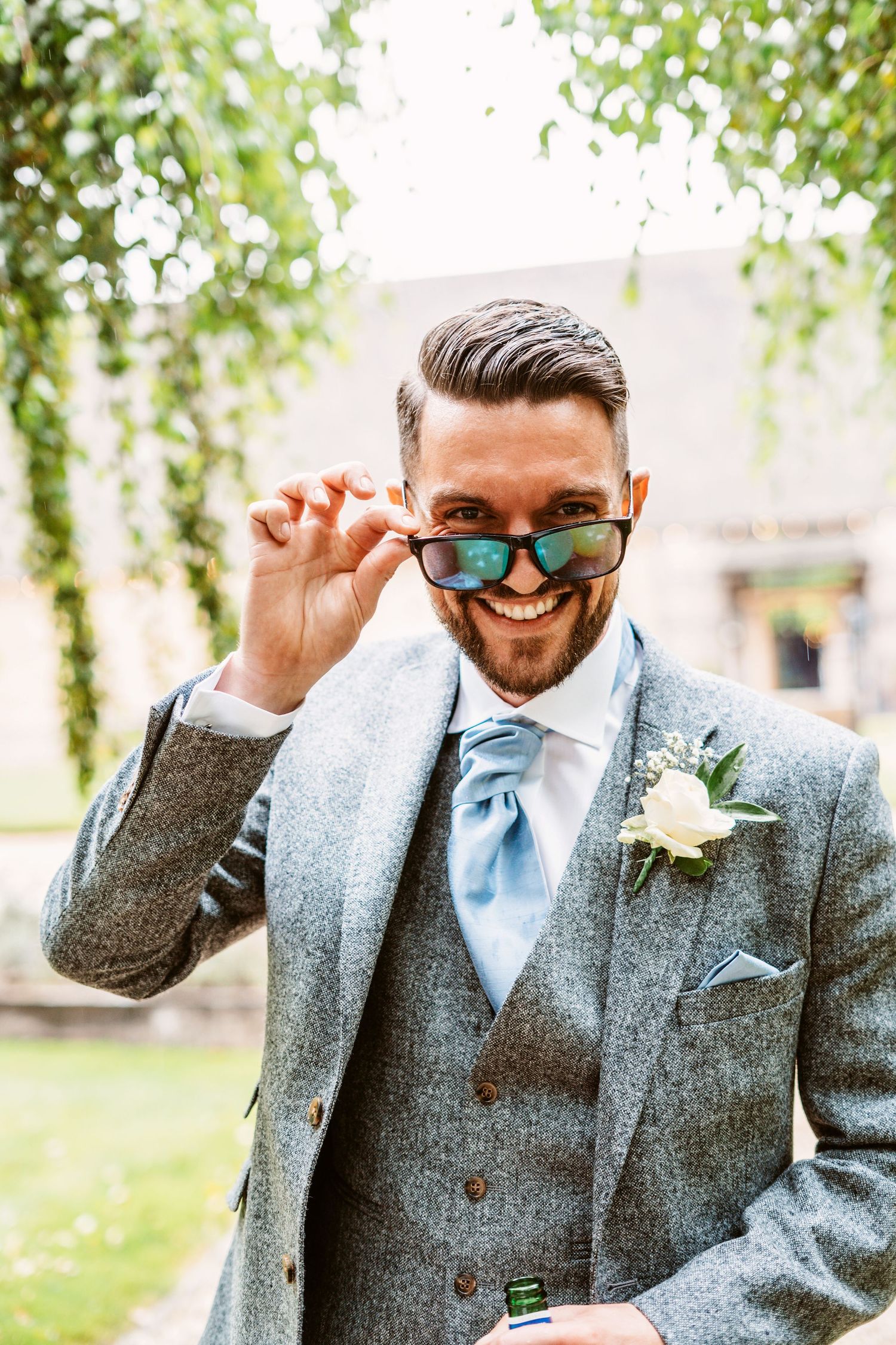 A joyful groom in a grey suit and blue tie adjusts his sunglasses while smiling outdoors.
