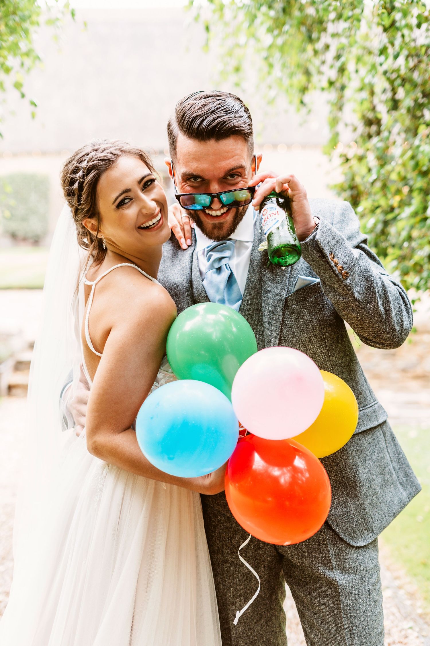A newlywed couple poses with colorful balloons while laughing together in a garden setting.