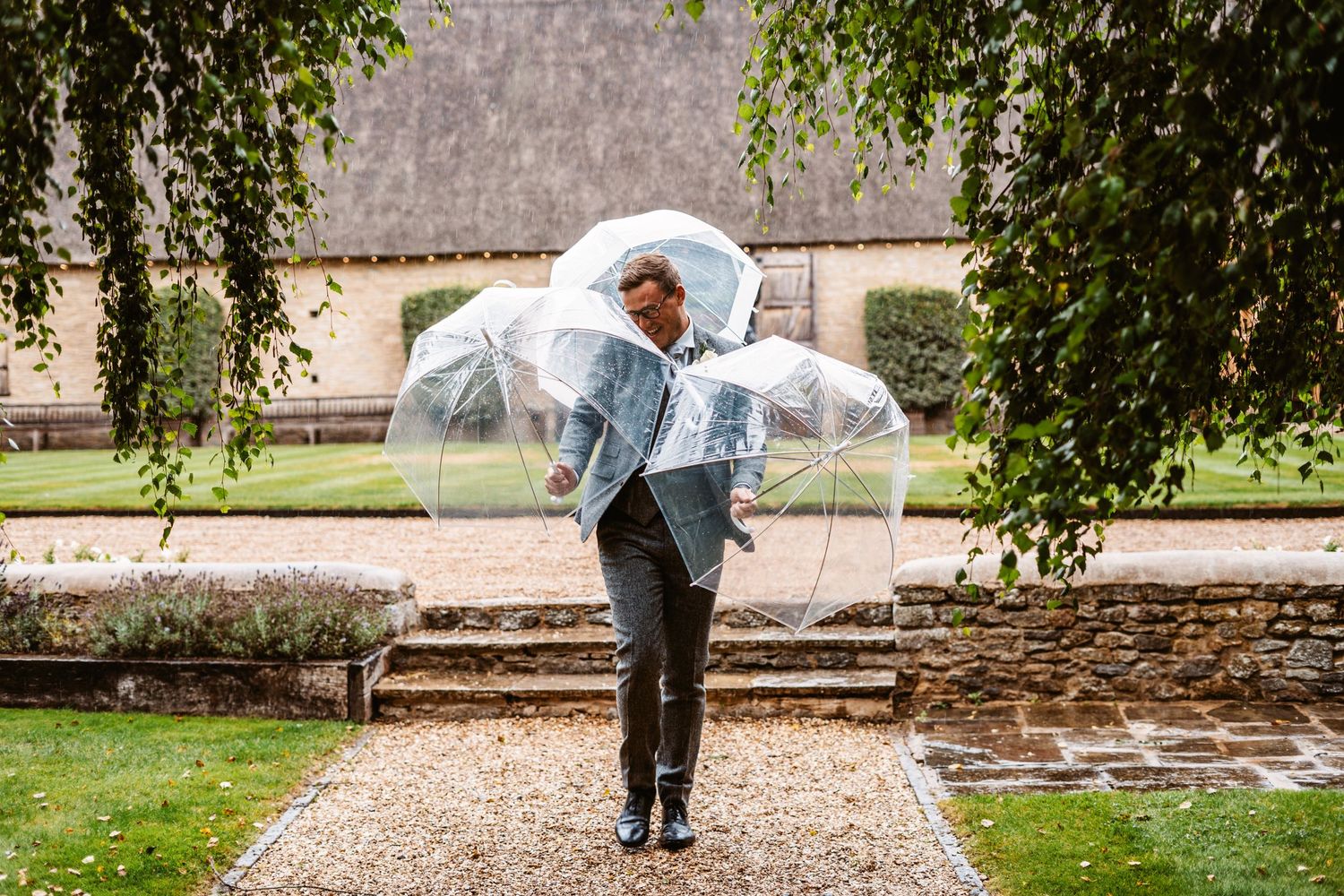 Person in formal attire holds clear umbrella while walking on stone path near historic building.