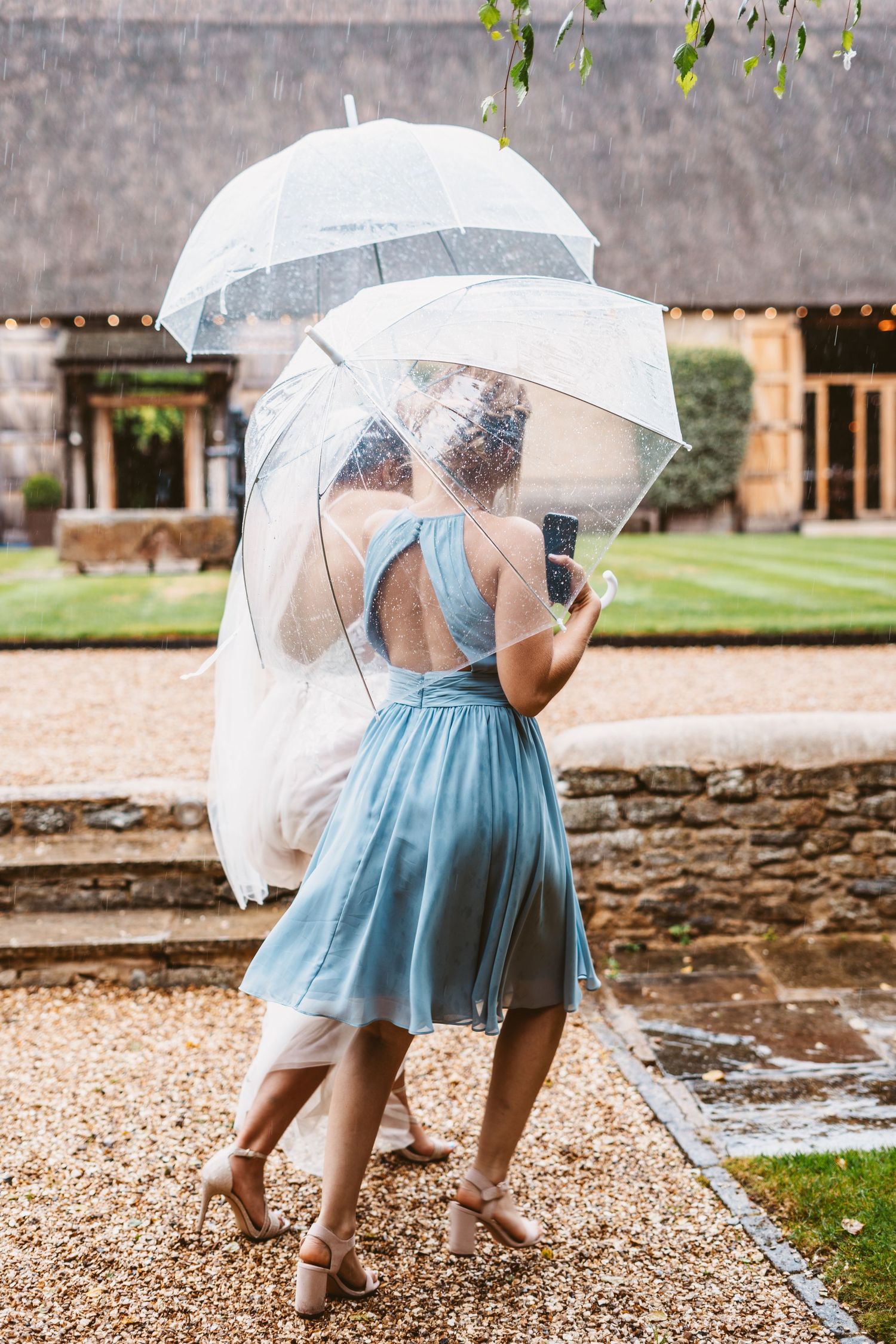 Two bridesmaids in blue dresses share clear umbrellas during a light rain.