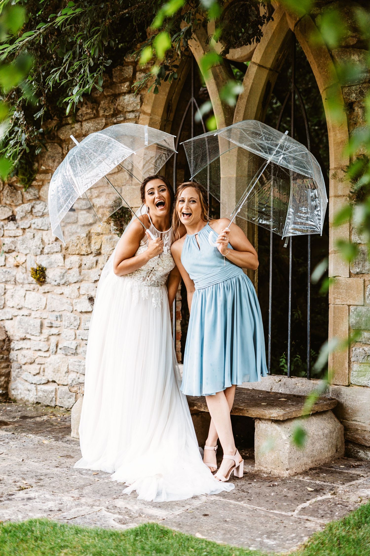 Two wedding attendees pose with clear umbrellas against stone wall backdrop.