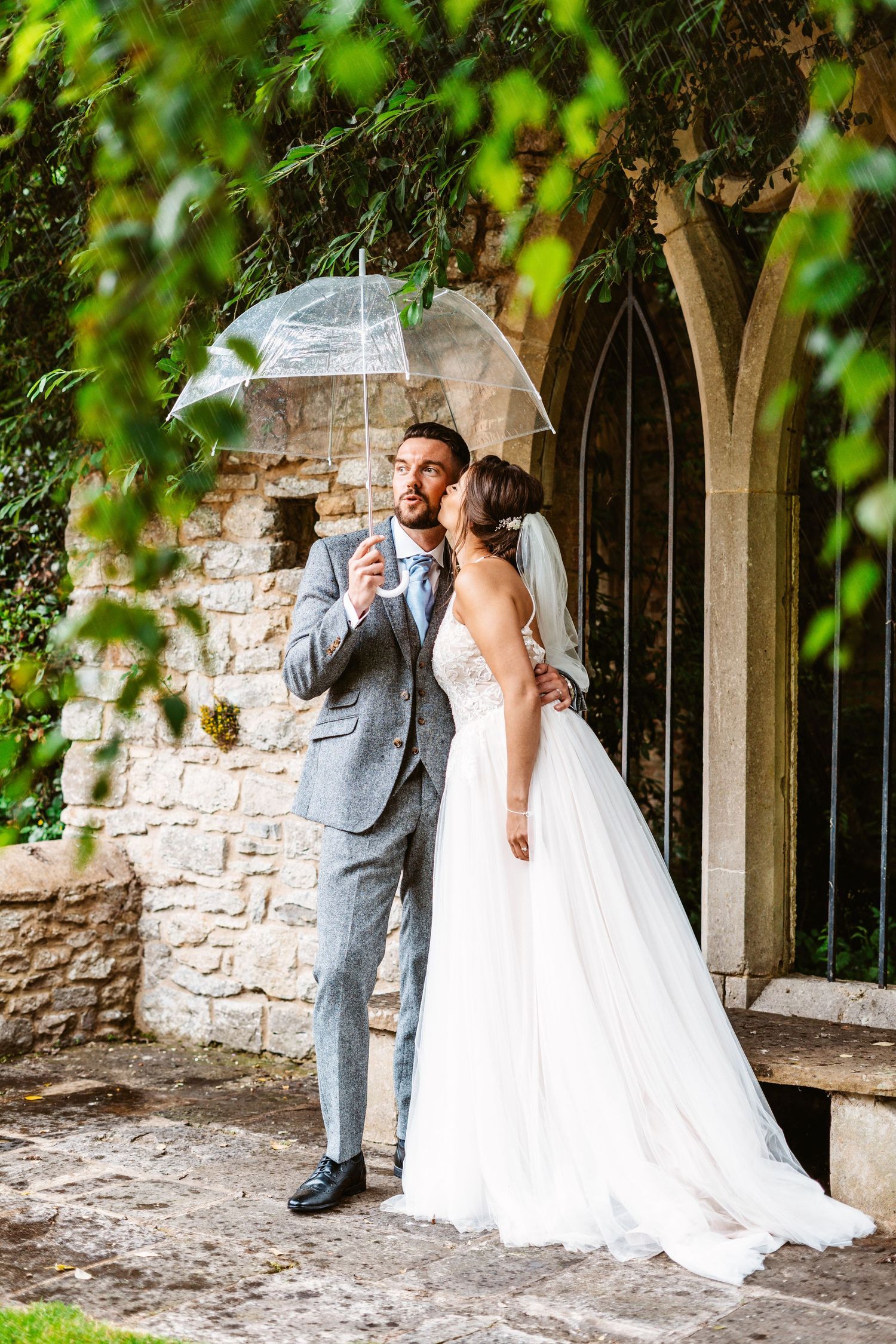 Wedding couple shares intimate moment under clear umbrella beside stone archway.