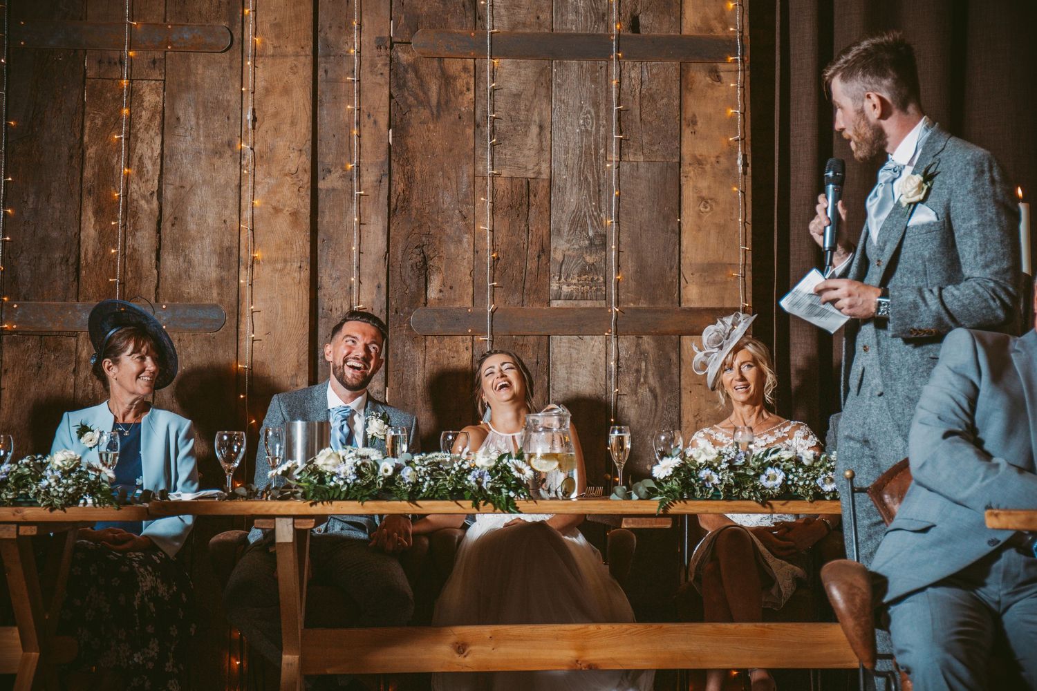 Wedding reception speech at a rustic wooden table with floral centerpieces and attentive guests.