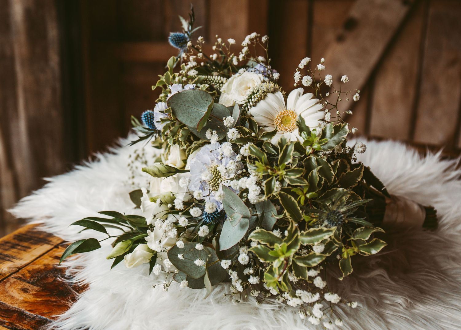 A rustic wedding bouquet with white daisies and eucalyptus leaves rests on a furry white surface.
