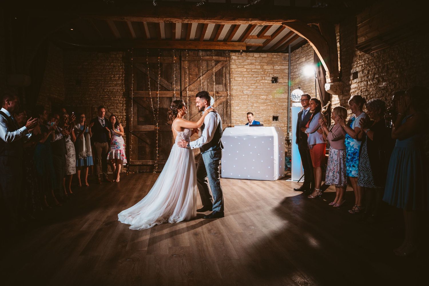 Romantic first dance on rustic wooden floor surrounded by guests.