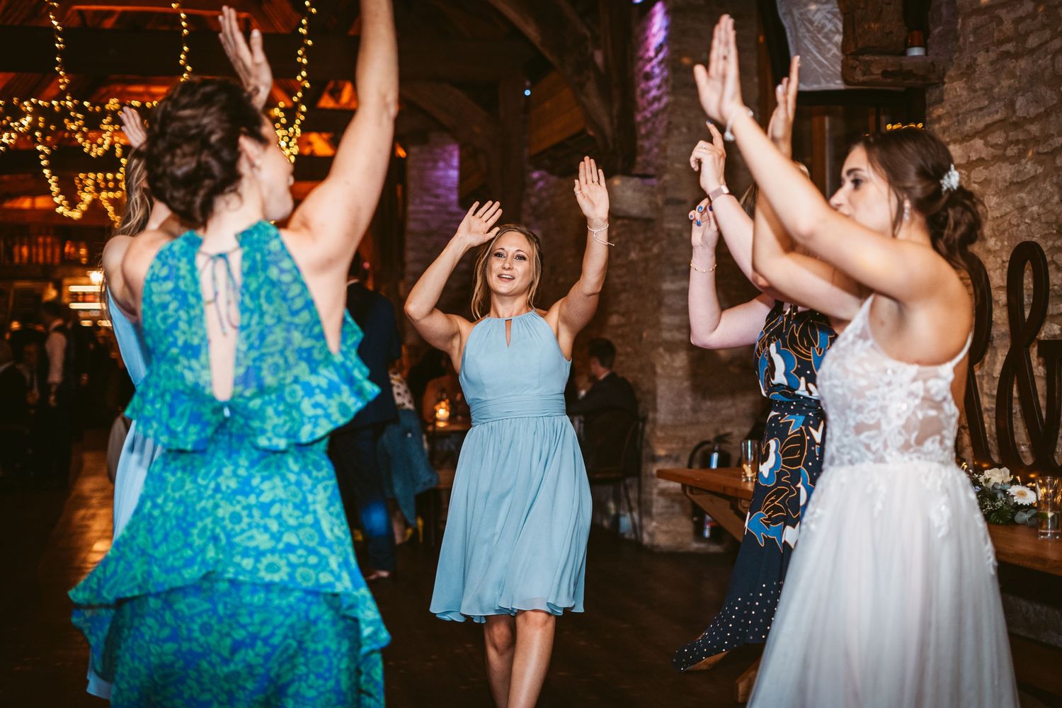 Wedding party dancing in a rustic barn venue with string lights and brick walls.