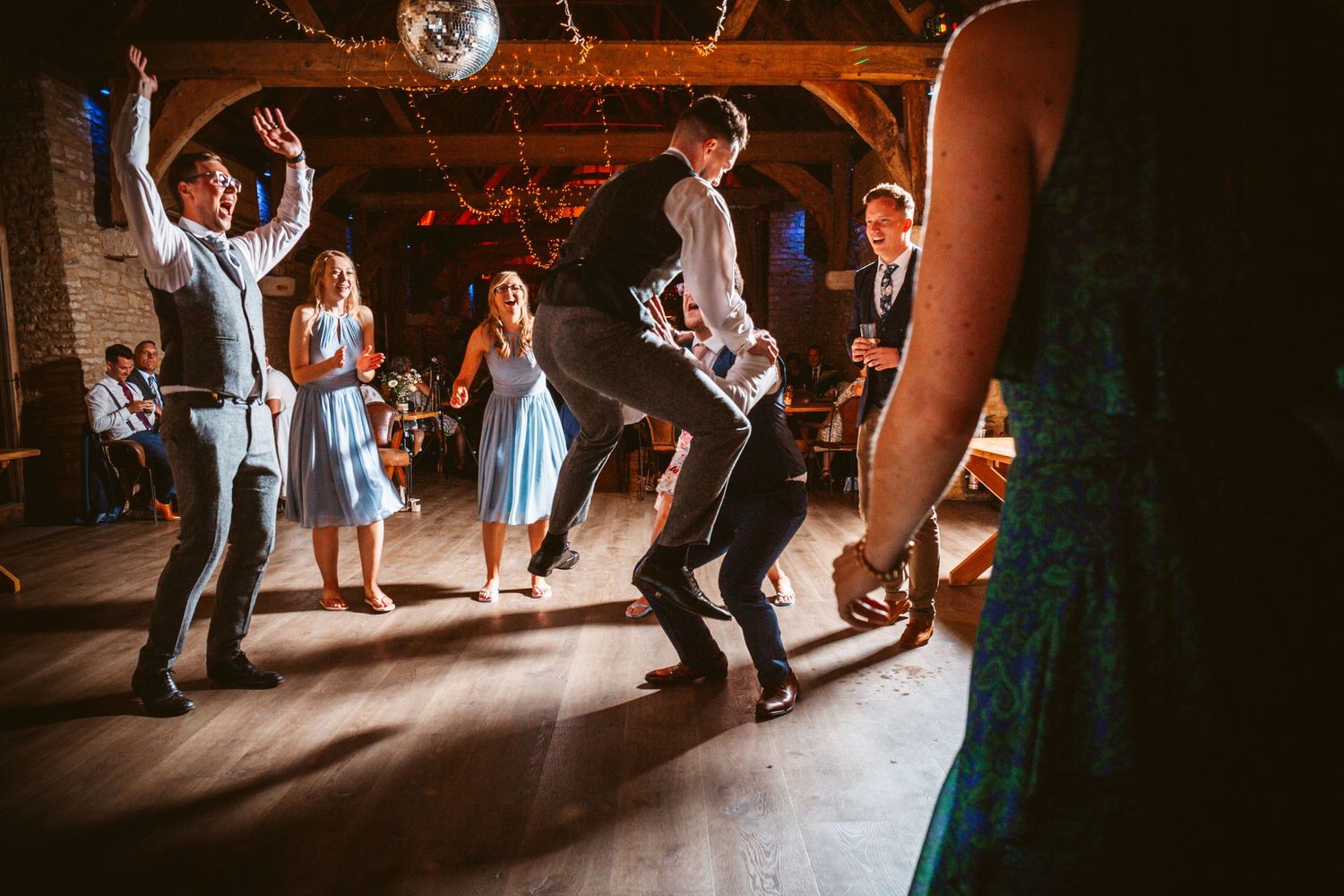 Wedding guests dancing under a disco ball in a dimly lit rustic barn setting.