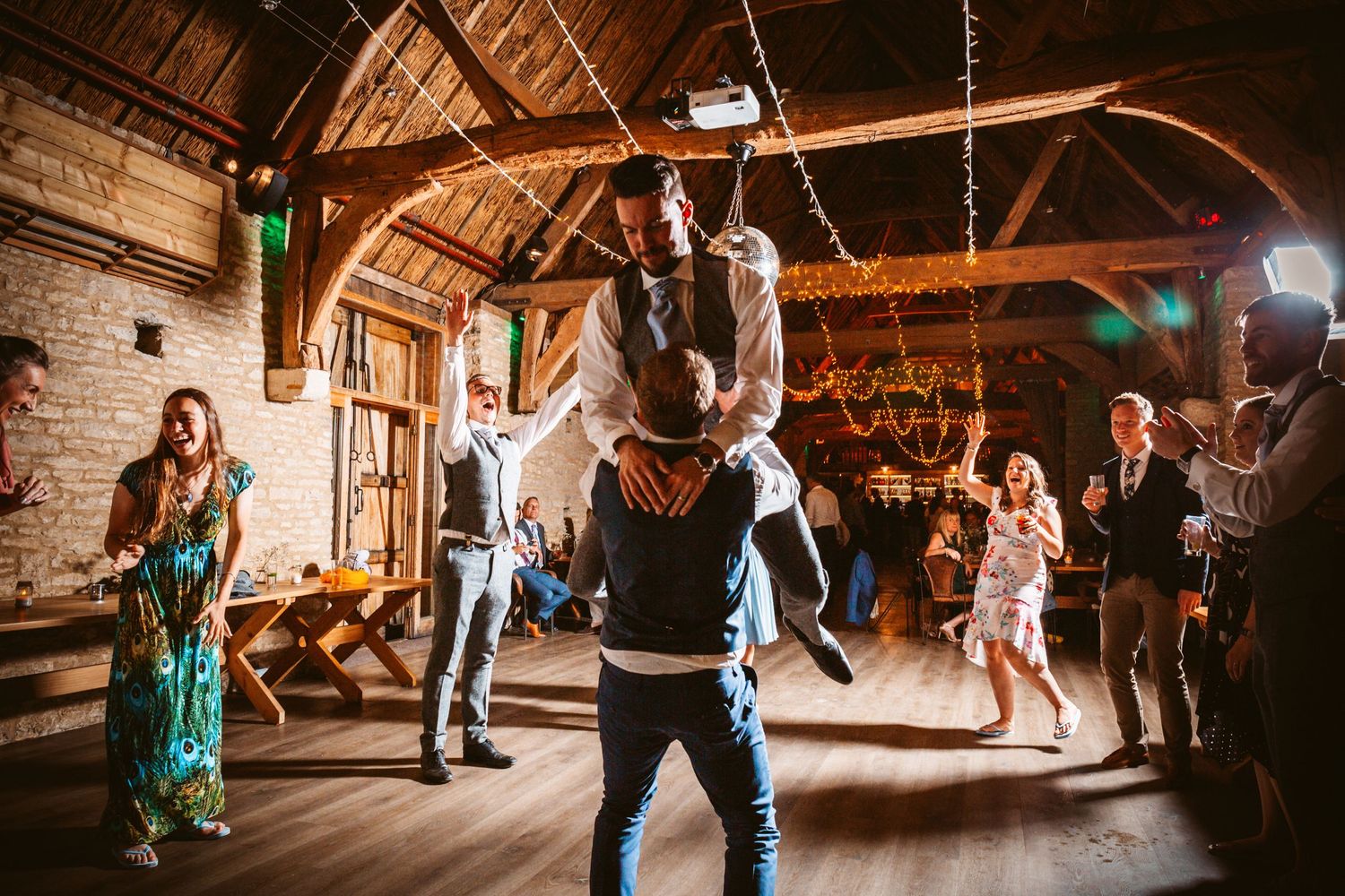 Guests celebrating and dancing at a barn wedding reception with wooden beams and string lights.