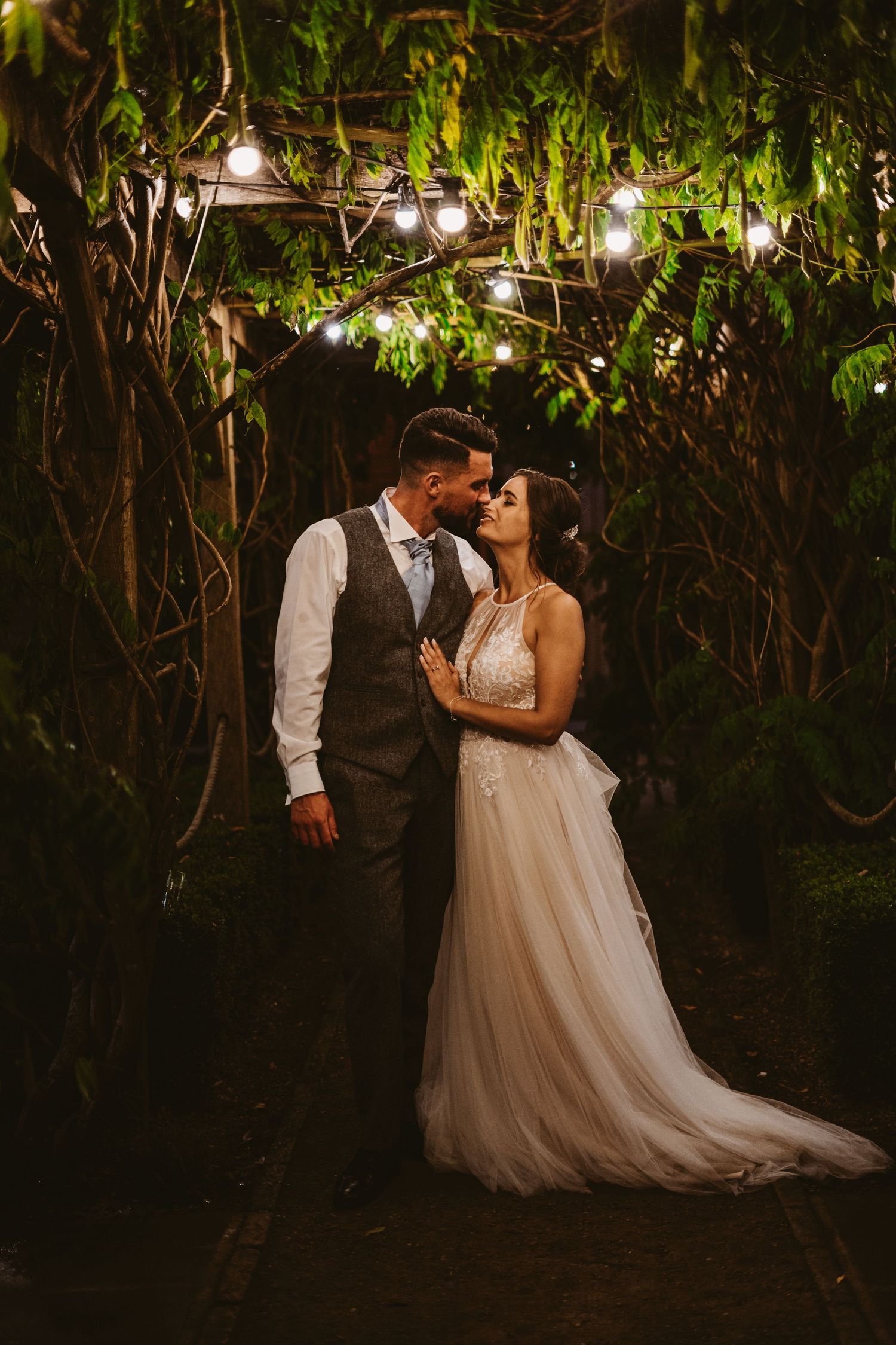 Romantic evening portrait of newlyweds under twinkling lights and lush garden foliage.