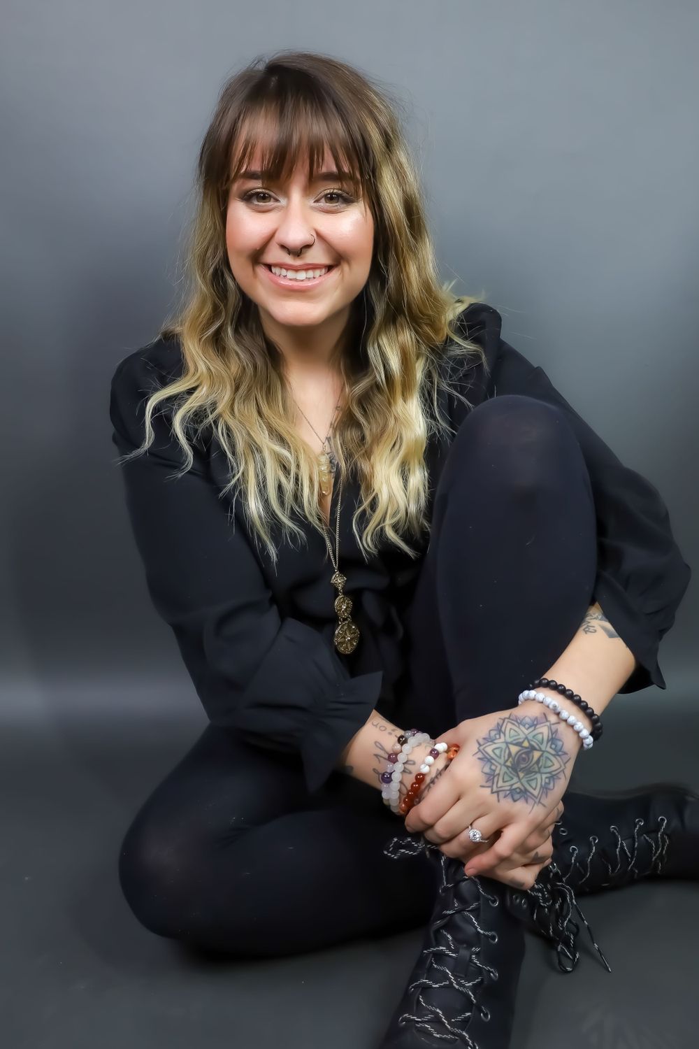 Studio portrait of person in black outfit showing tattoos and jewelry against dark background.