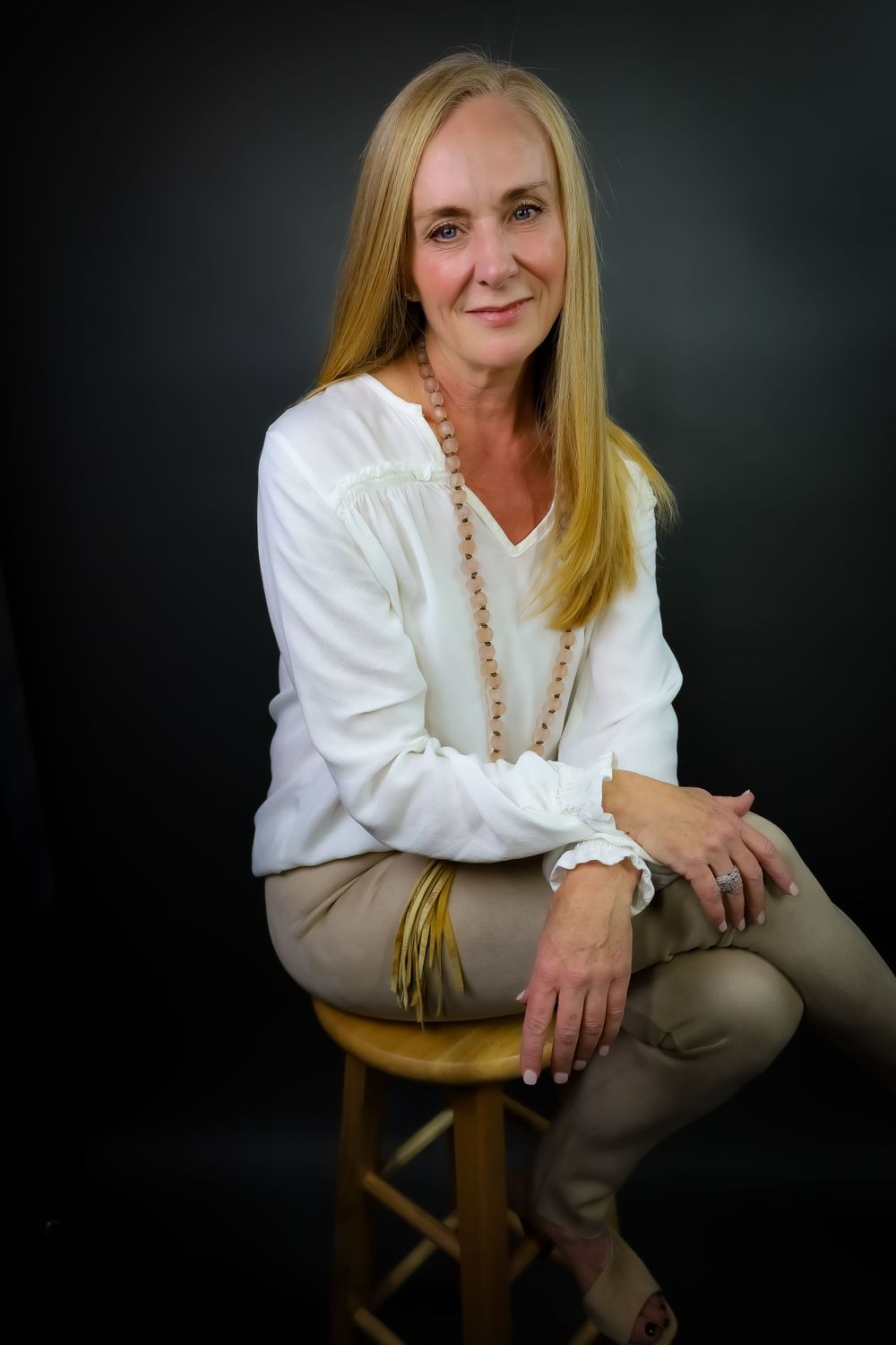 Elegant portrait of person in white blouse and beige pants seated on wooden stool.