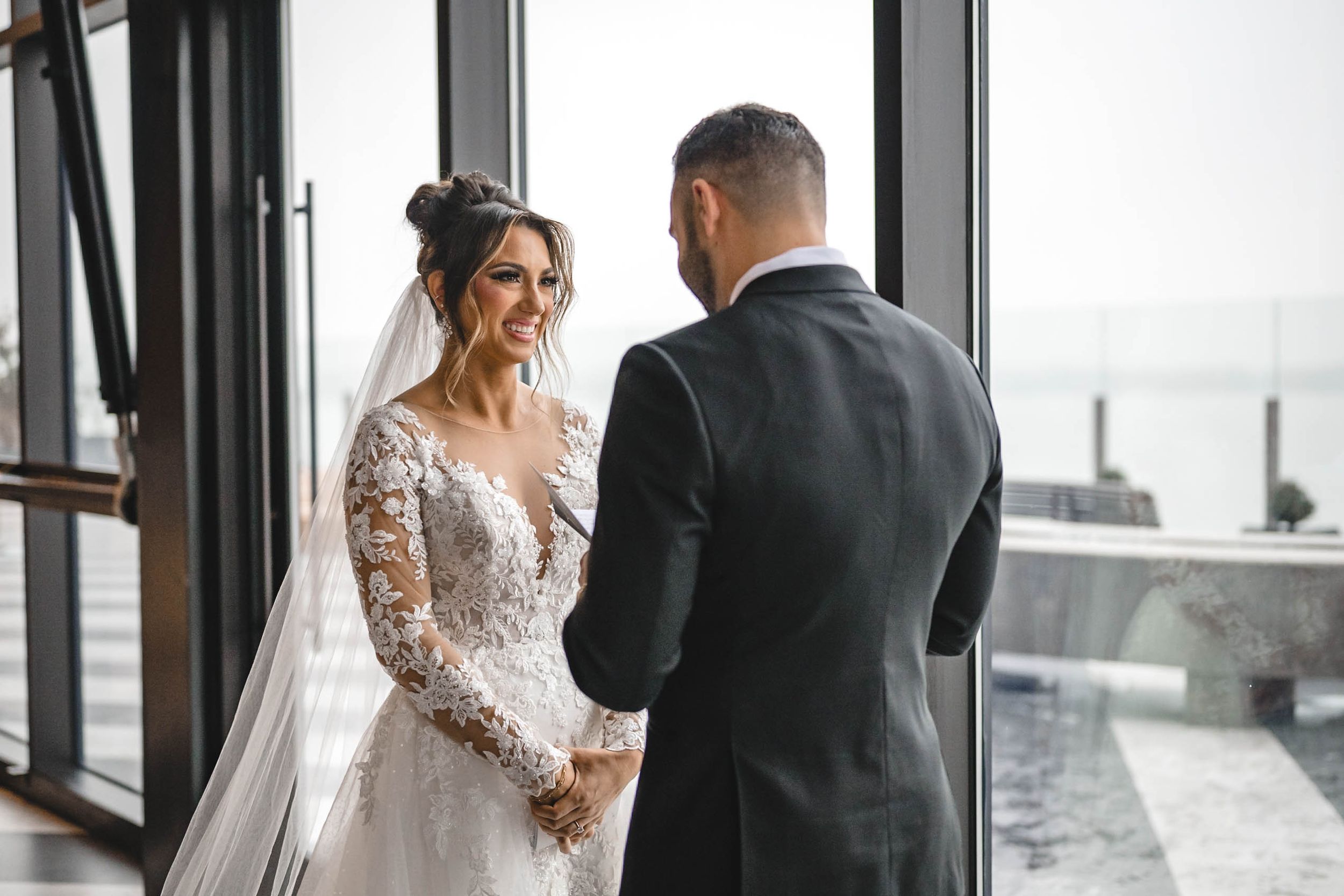 Bride in long-sleeved lace gown and groom sharing private moment by window with city view.
