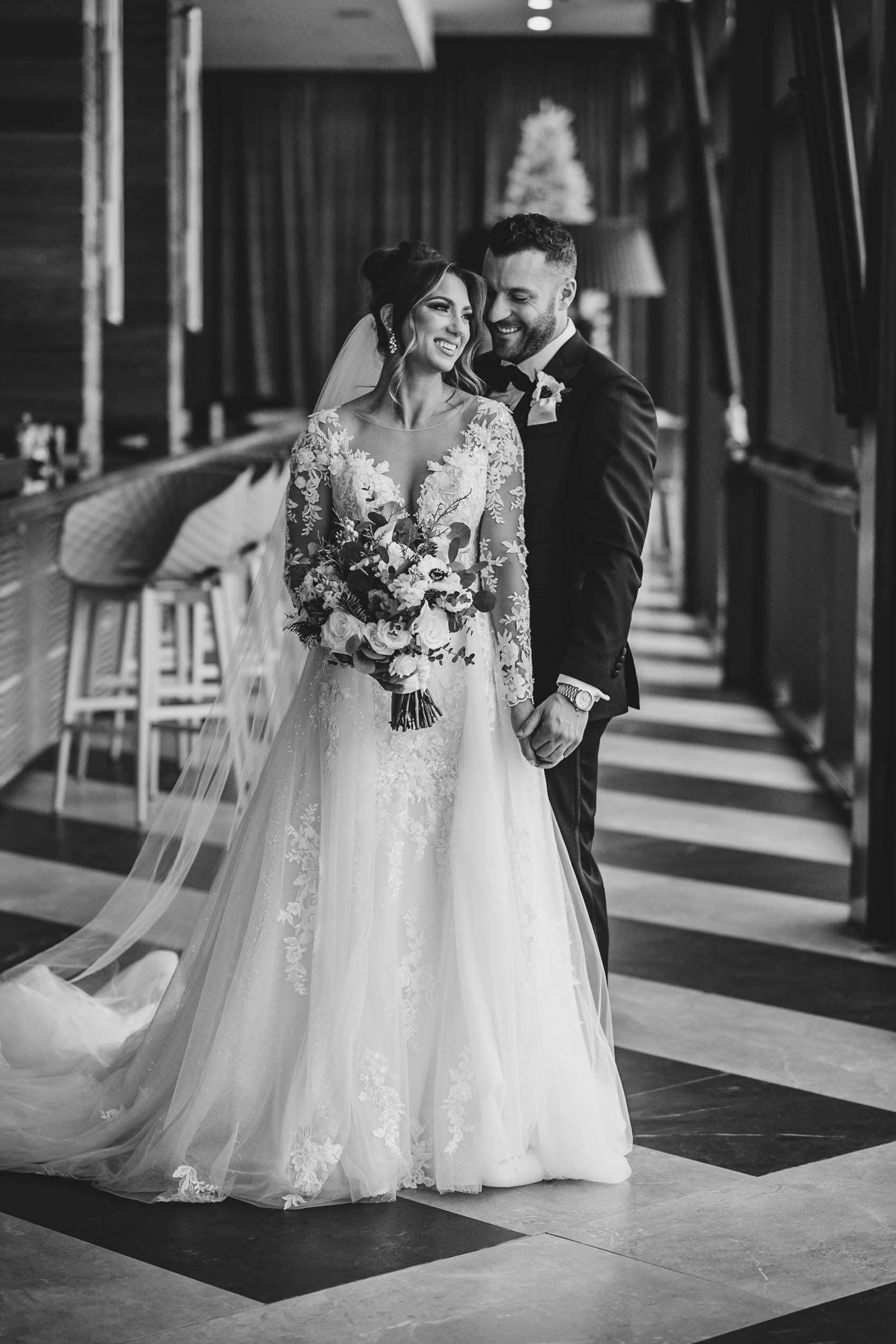 A romantic wedding couple poses together in an elegant hallway with a flowing white dress and bouquet.