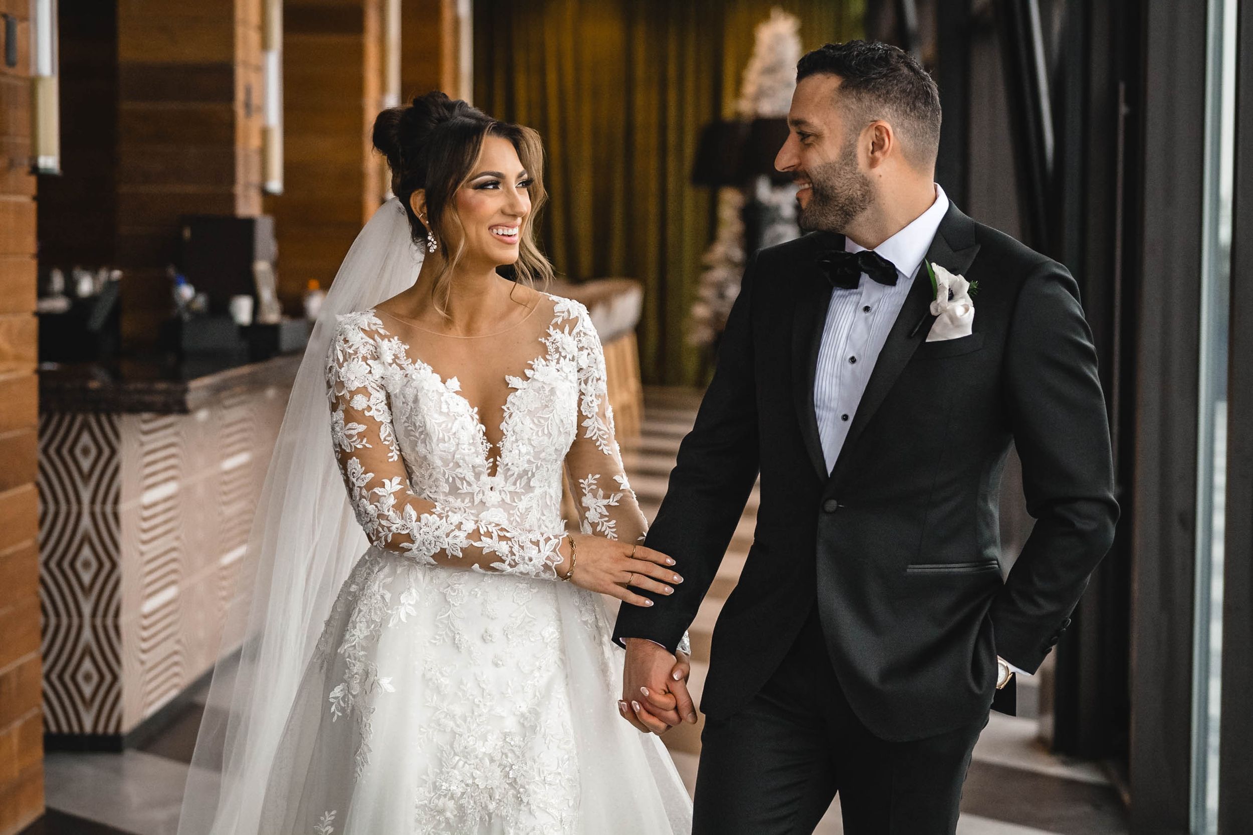 Elegant wedding couple walking together in formal attire through a modern venue hallway.