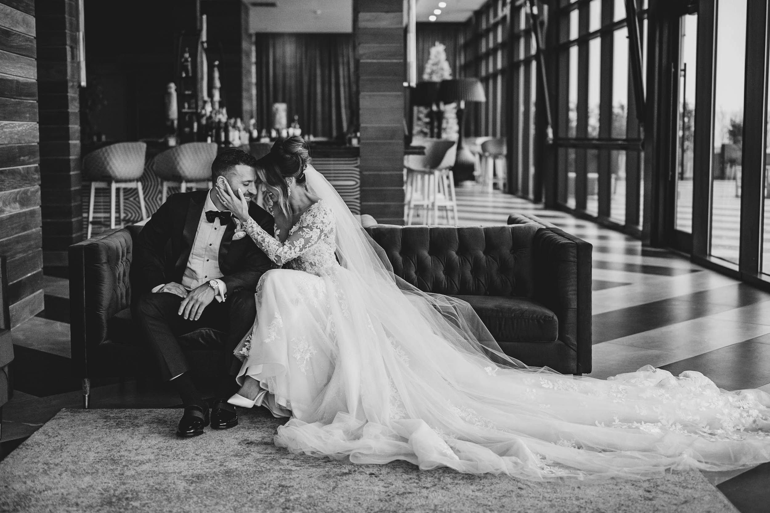 Bride in flowing white gown poses with partner in contemporary venue with dramatic lighting.