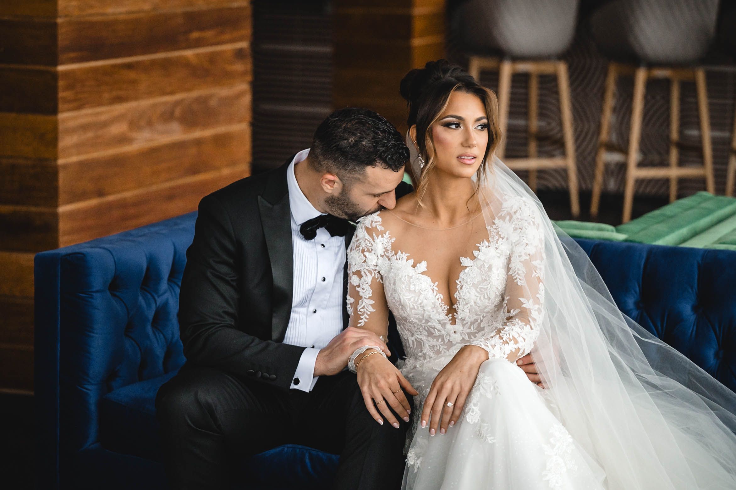 Couple shares tender moment on blue velvet seating in rustic-modern wedding venue.