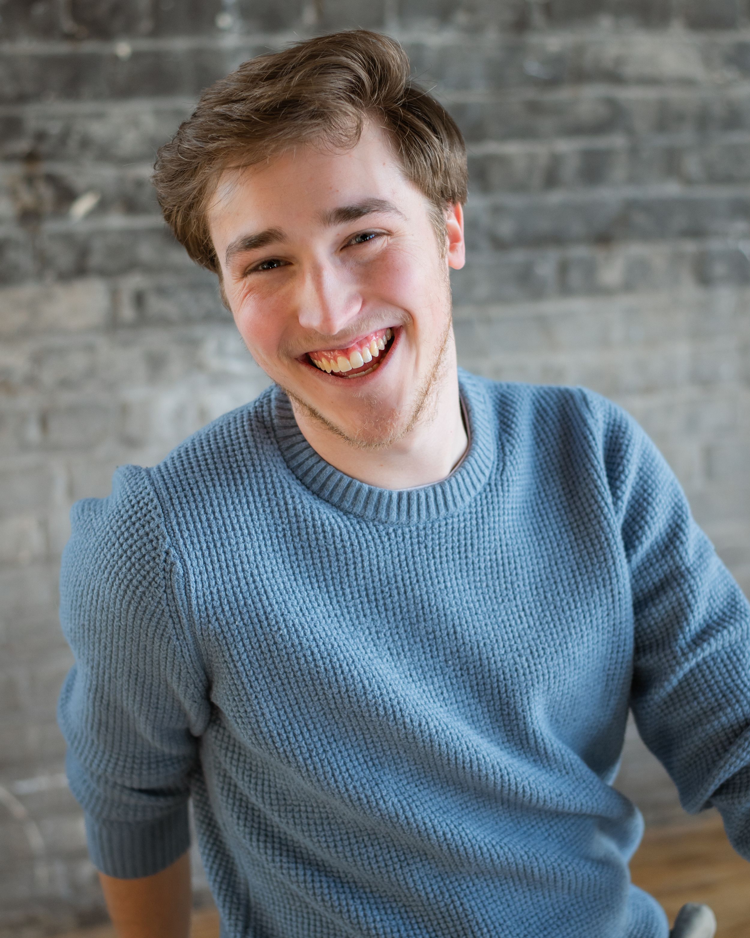 Portrait of someone wearing a blue sweater smiling warmly against a brick wall.