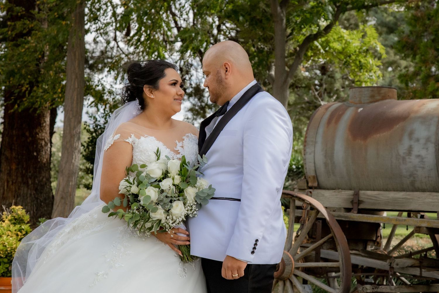 Bride and groom share a romantic moment holding a white rose bouquet near a rustic vintage wagon.