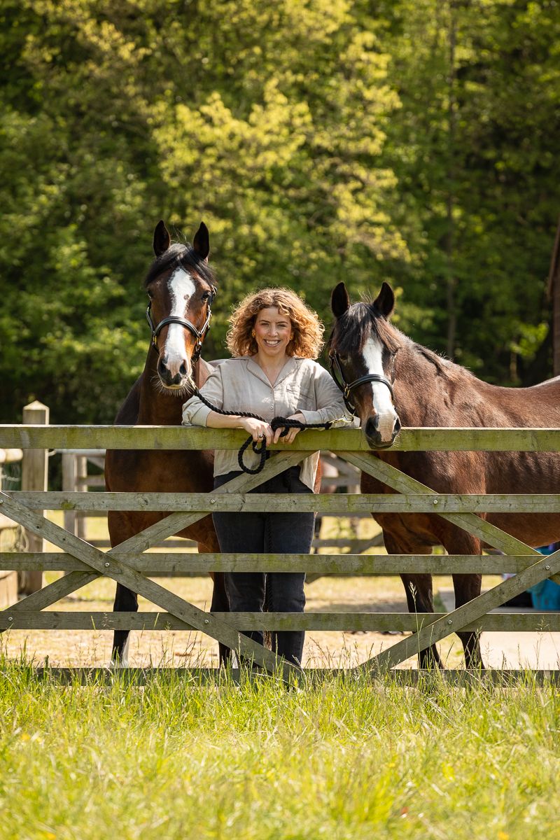 A woman leans on a wooden fence with two horses in a pastoral setting.