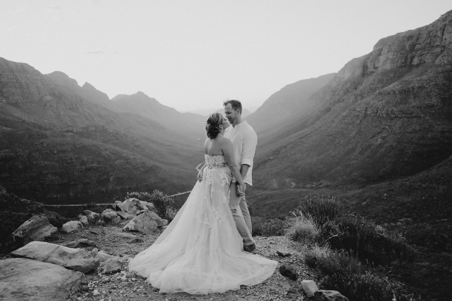 A wedding couple stands together overlooking a dramatic mountain valley in black and white.