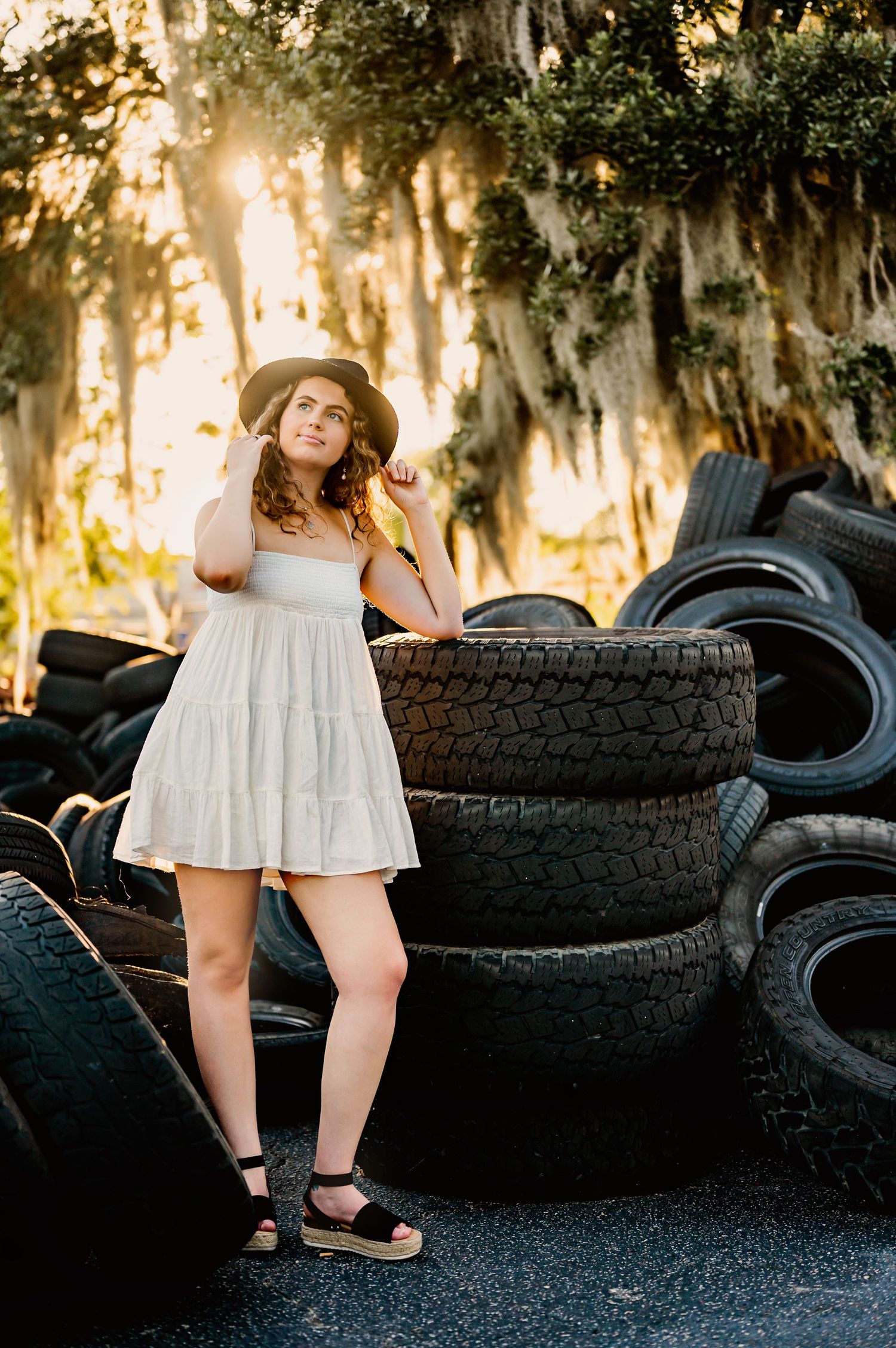 Artistic portrait with woman in white dress posed against stacked tires at sunset.