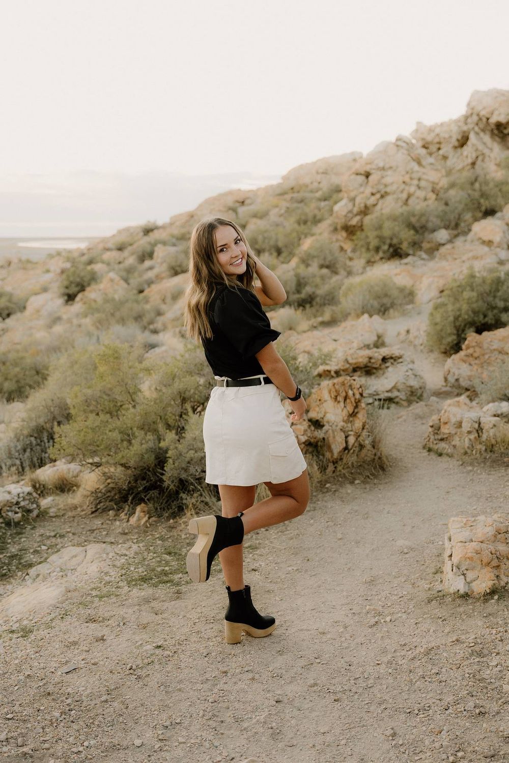 Fashion portrait featuring black top, white skirt and boots on mountain path at golden hour.
