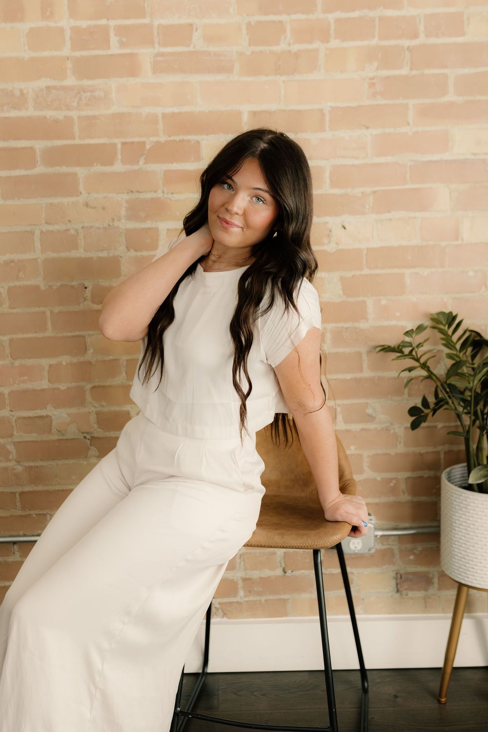 Woman in elegant white dress sits on chair by brick wall with potted plant.