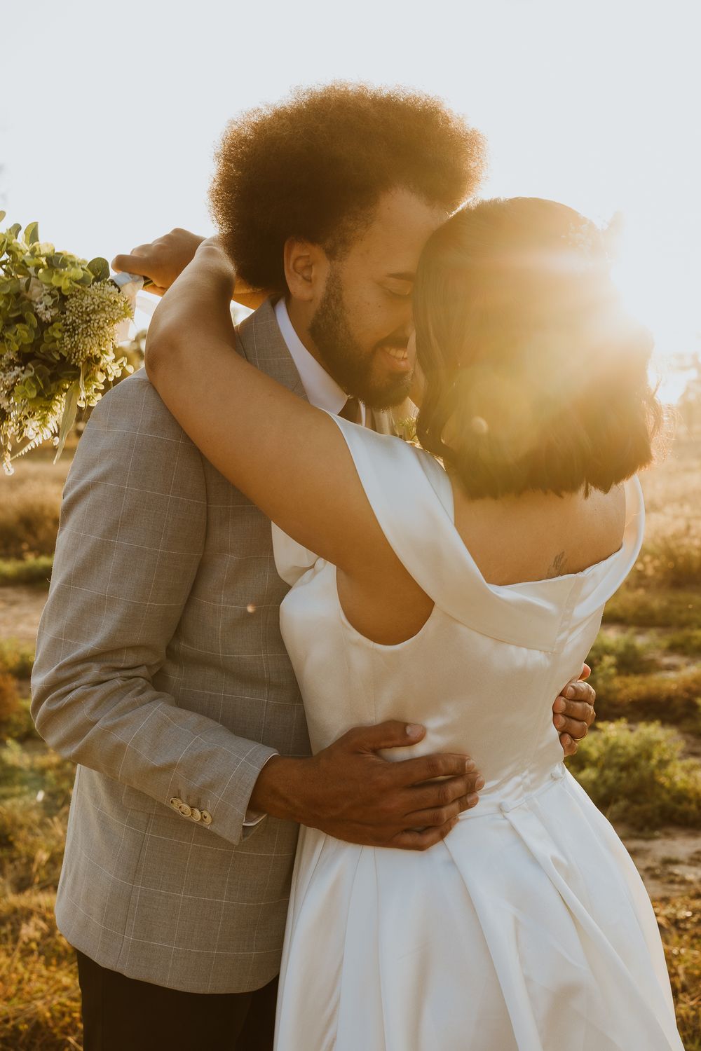 Newlywed couple holding bouquet while sharing intimate embrace in warm sunset glow in open countryside field.