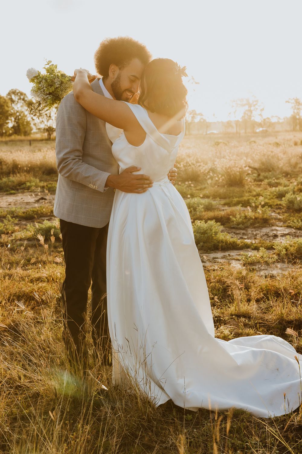 Bride and groom embracing in golden sunset light in rustic field, bride wearing elegant white off-shoulder gown.