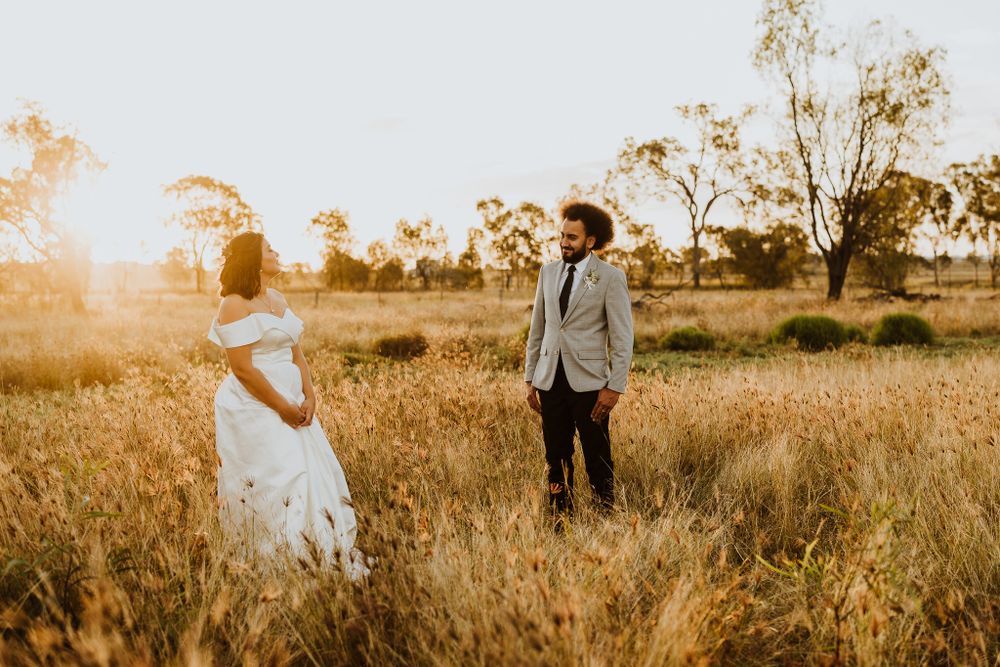 Bride and groom standing apart in sunlit golden wheat field, sharing a romantic glance at sunset.