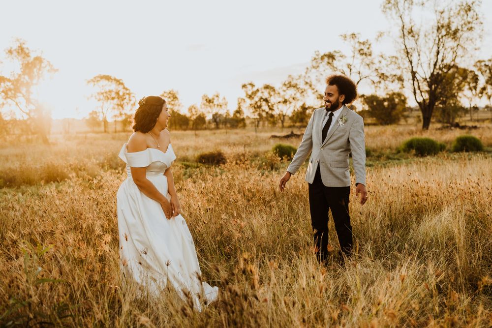Bride in white off-shoulder gown and groom in grey suit walking through golden sunlit field at sunset.