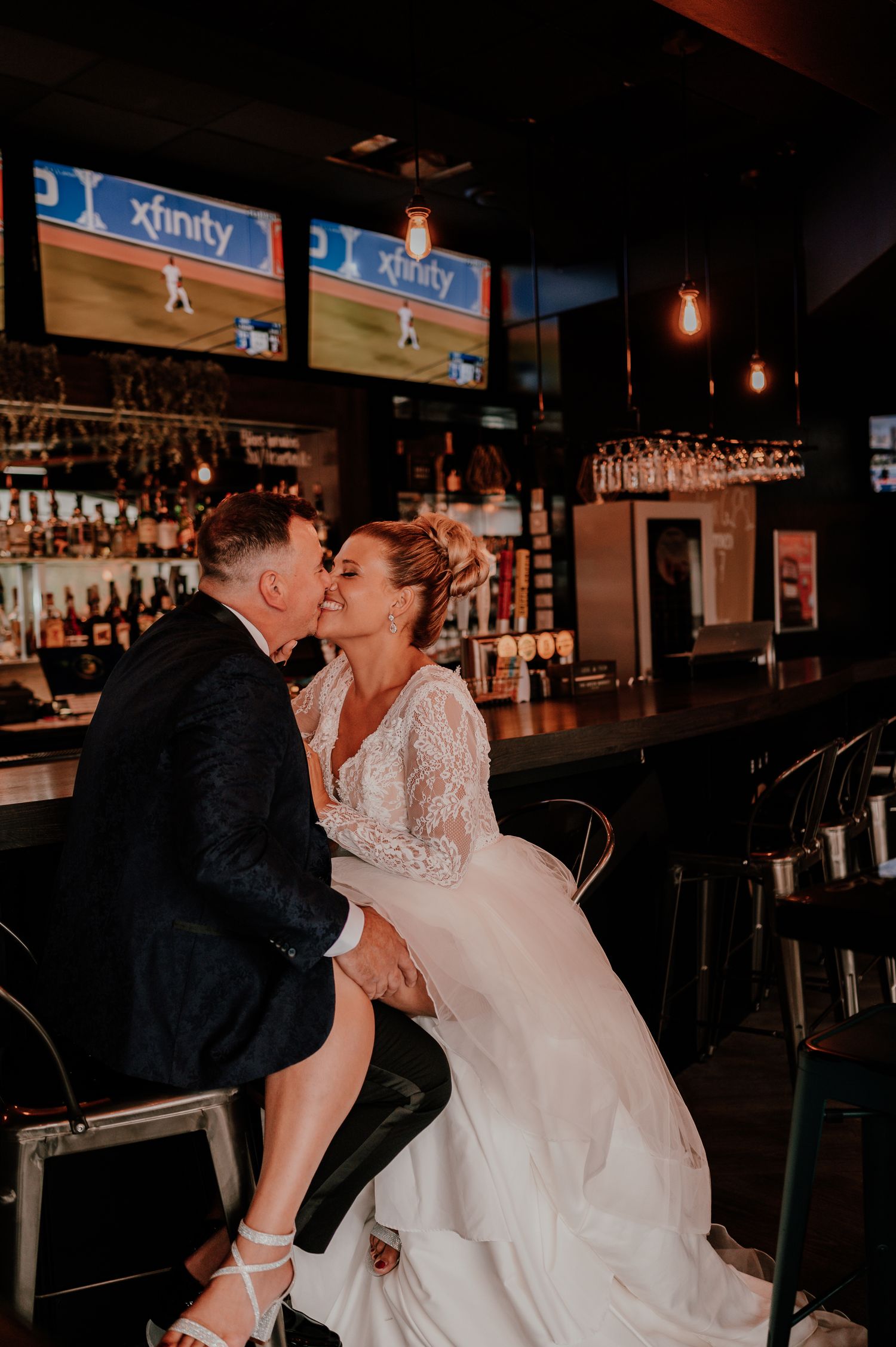 A couple shares a romantic kiss at a sports bar during their wedding celebration, with TVs and liquor bottles visible behind them.