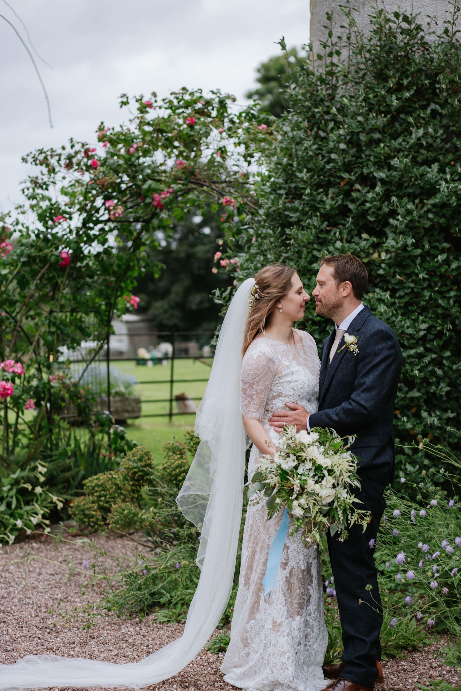 A wedding couple poses together in a garden with blooming roses and a flowing white veil.