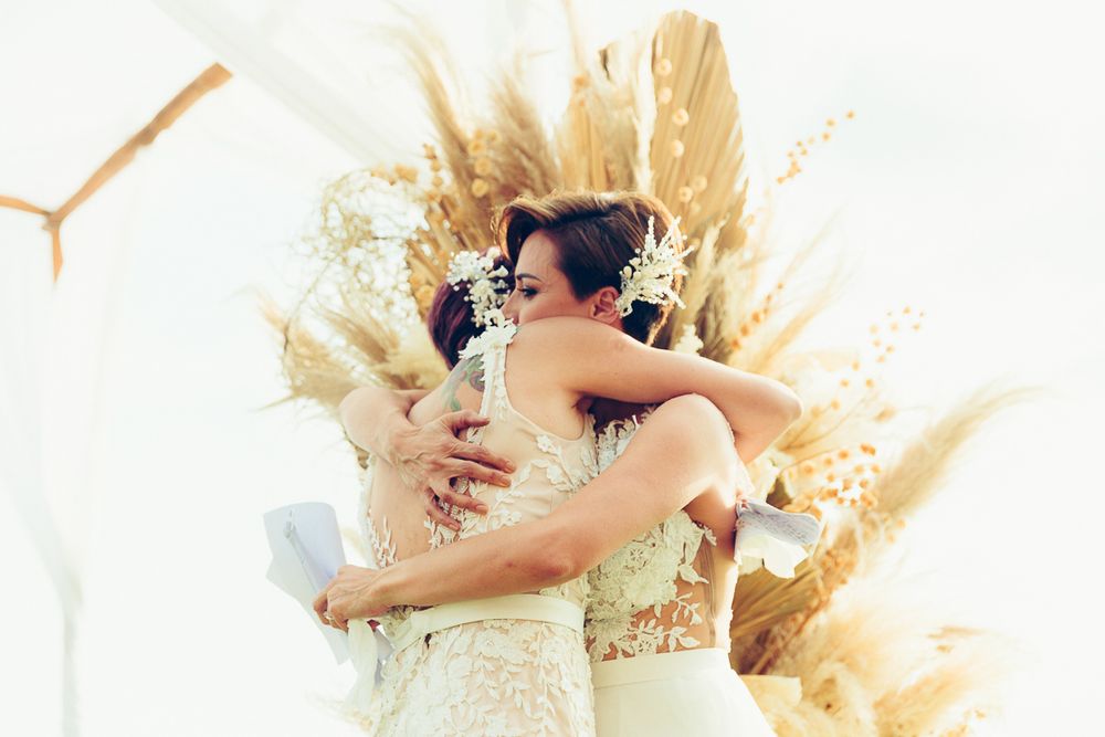 Couple embracing in white wedding attire against a backdrop of dried pampas grass decorations.