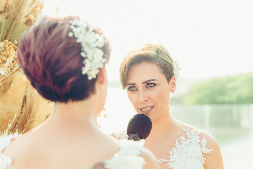 Two people in white wedding attire share an intimate moment while speaking into a microphone outdoors.
