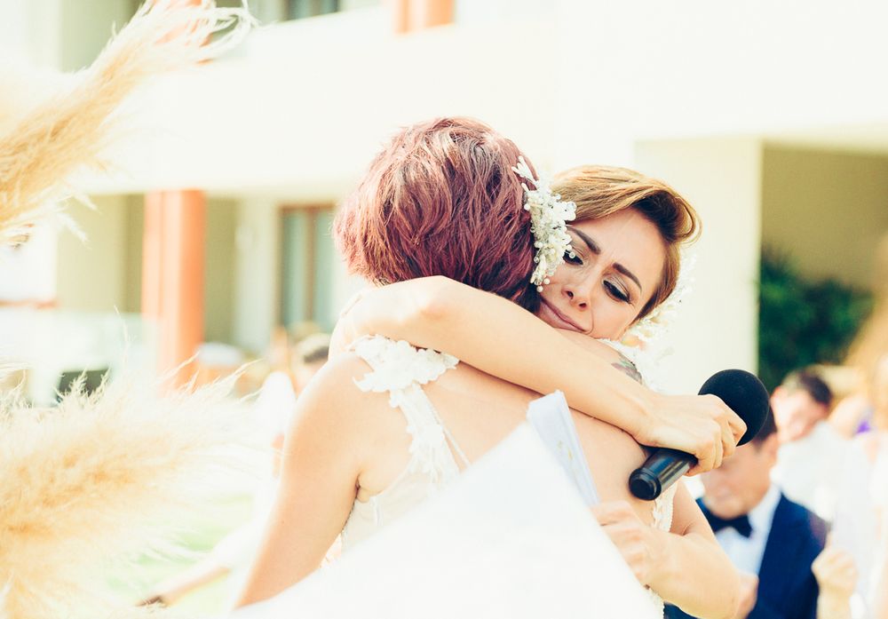 Two people in white wedding dresses embrace during an outdoor celebration.